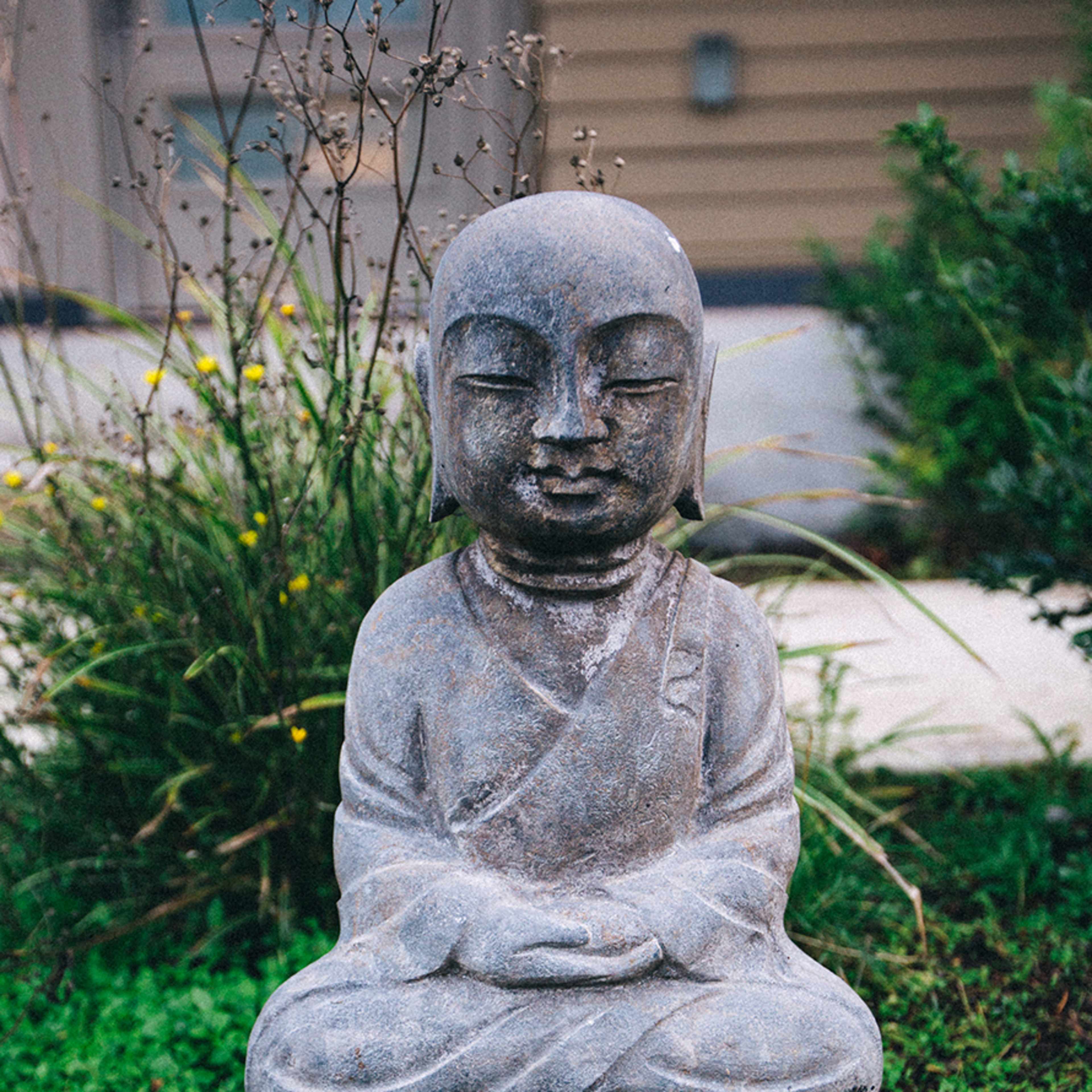 A stone statue of a seated Buddha is surrounded by greenery and small stones in a garden setting.