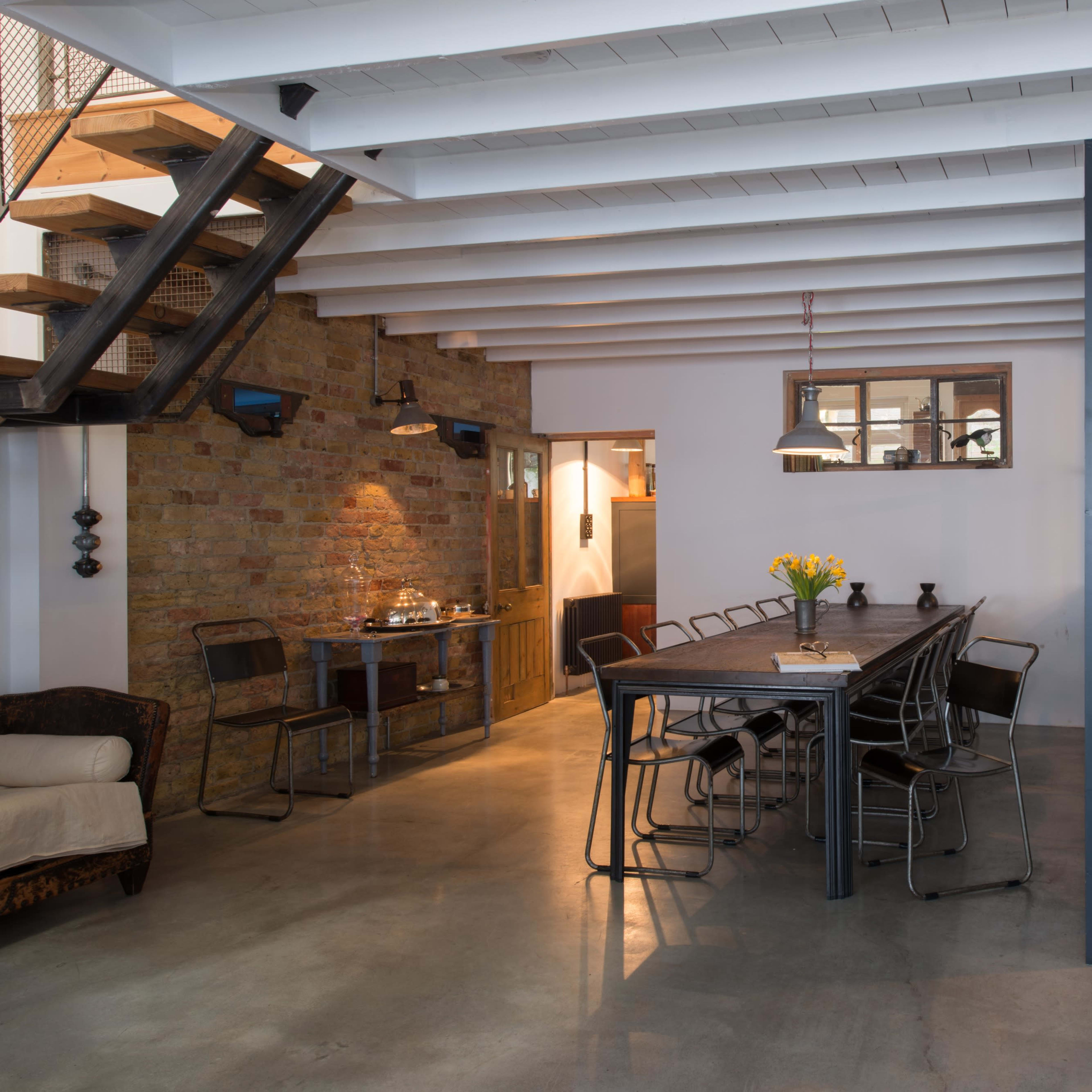 A modern, open-concept interior space featuring a dining table surrounded by chairs, a staircase, and a mix of brick and white walls.