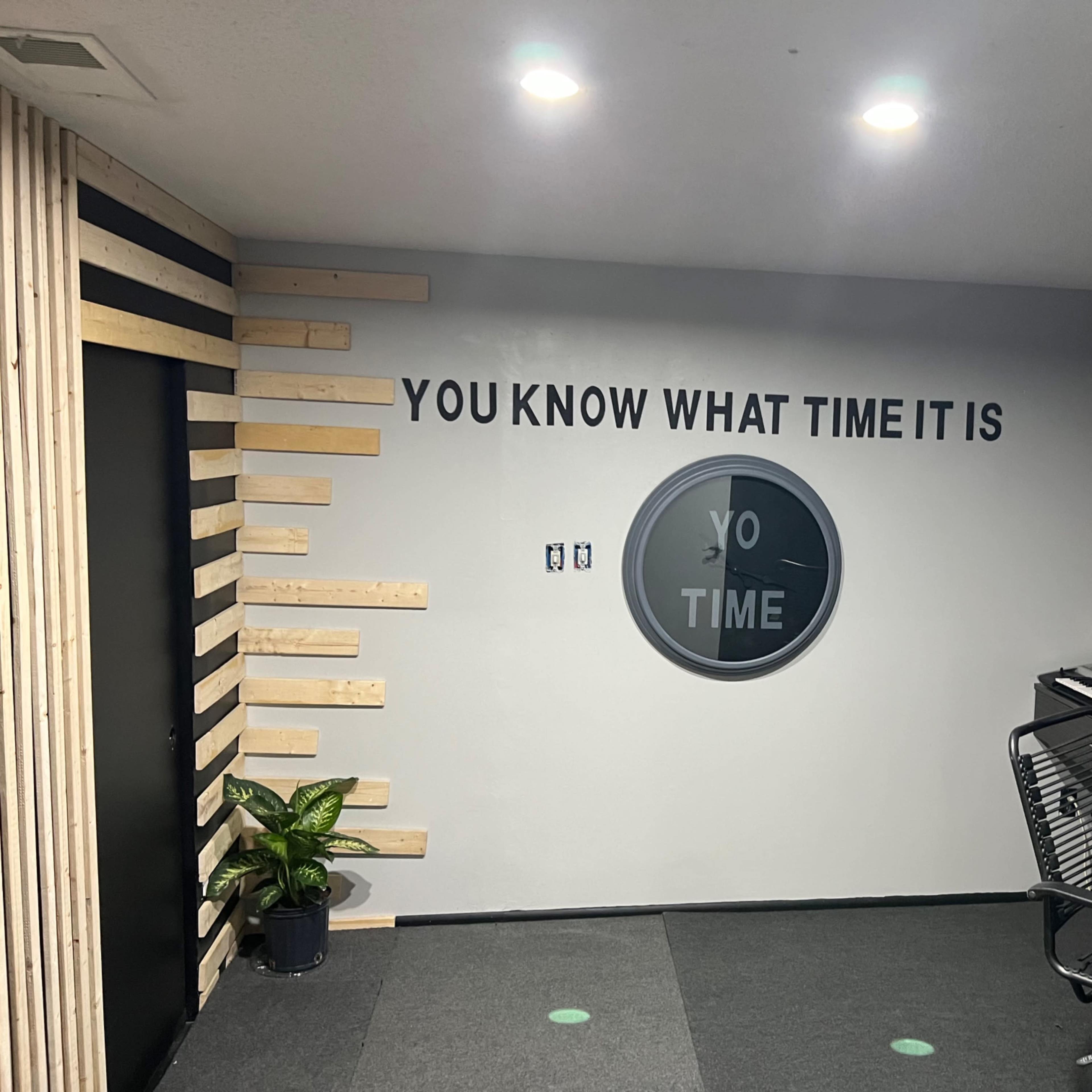 A modern office space with a wall featuring the phrase "YOU KNOW WHAT TIME IT IS" and a circular clock design, accompanied by a plant and a desk with a keyboard.