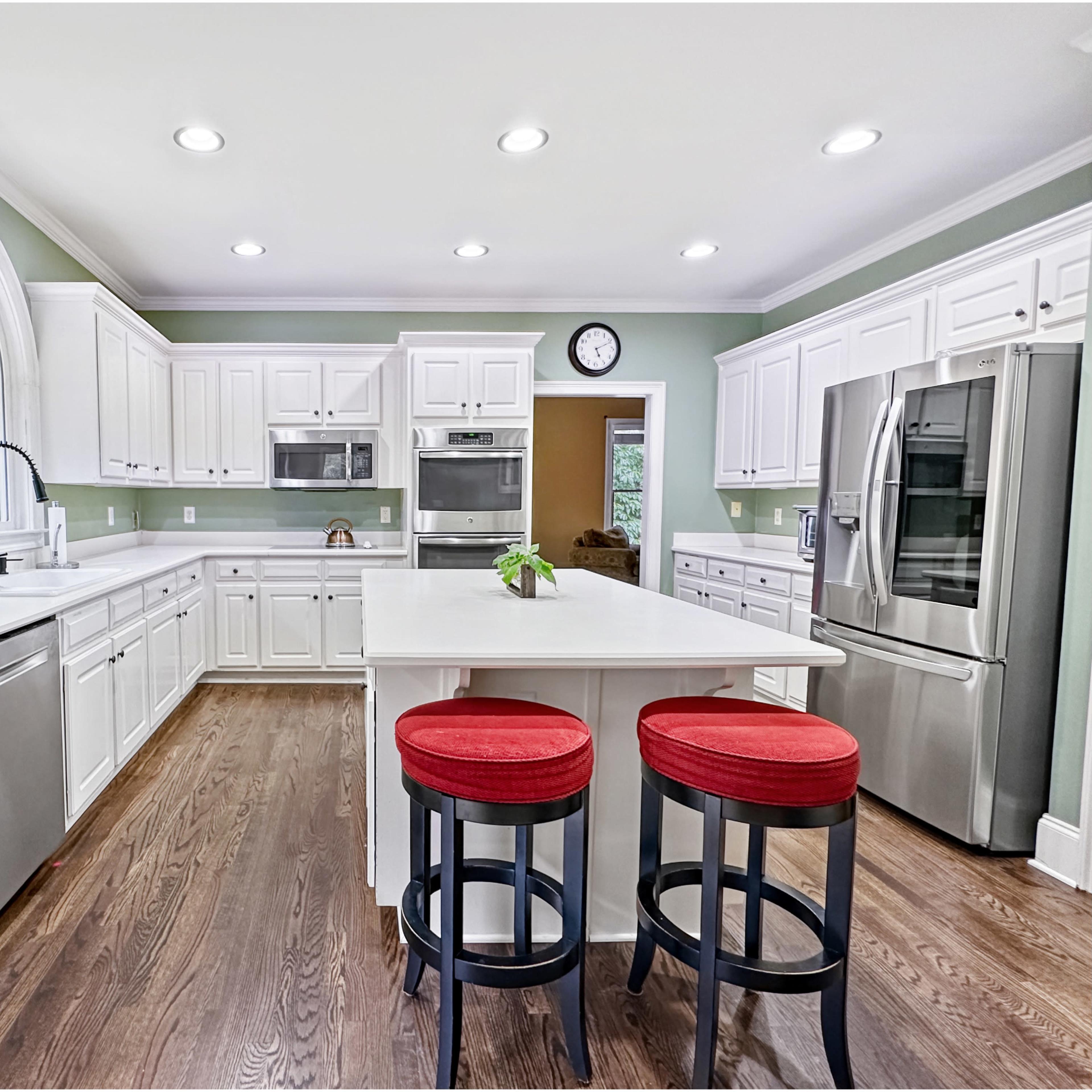 The kitchen features white cabinetry, stainless steel appliances, a central island with red stools, and natural light from a large window.