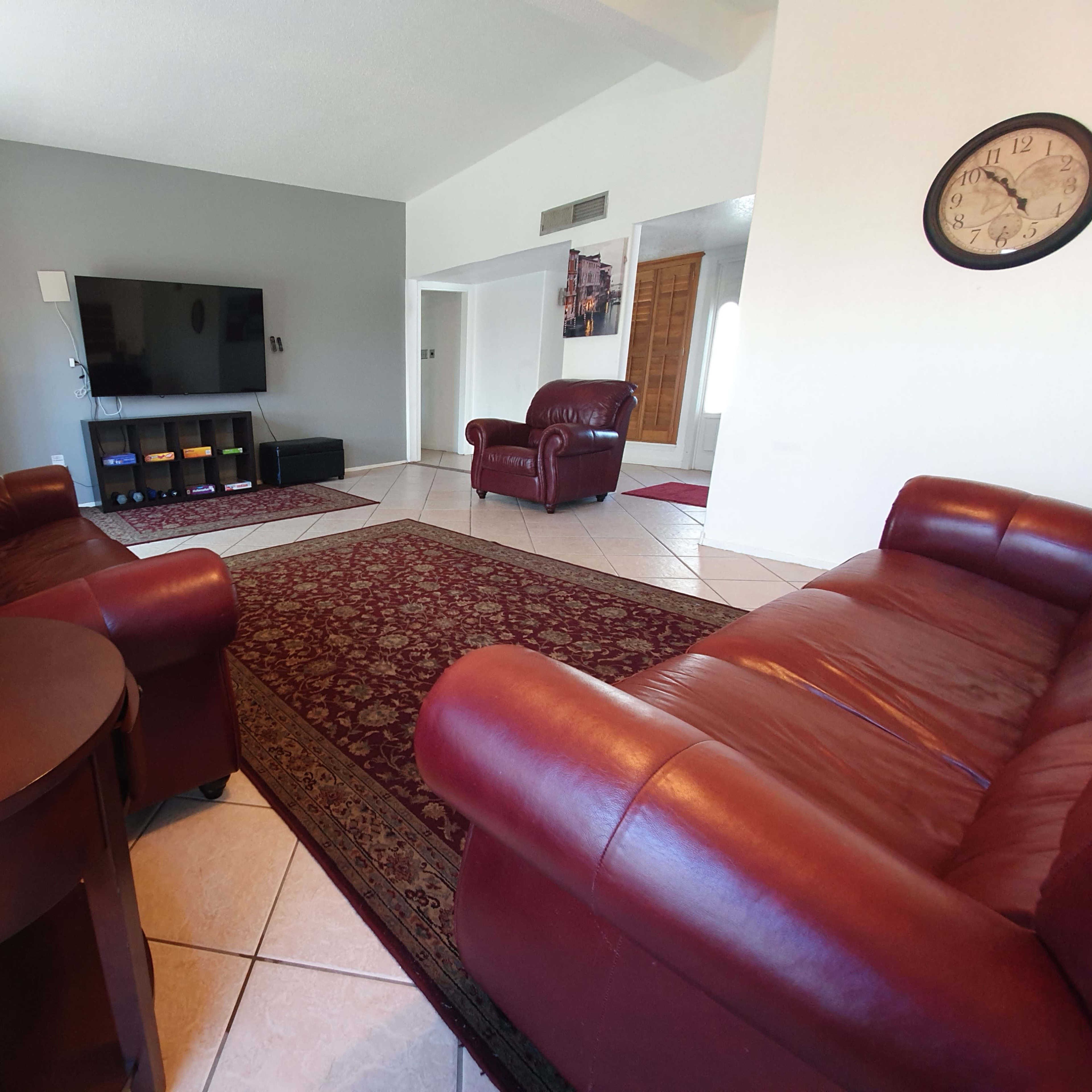 A living room features two red leather chairs and a matching sofa, with a large television on the wall opposite a patterned rug.