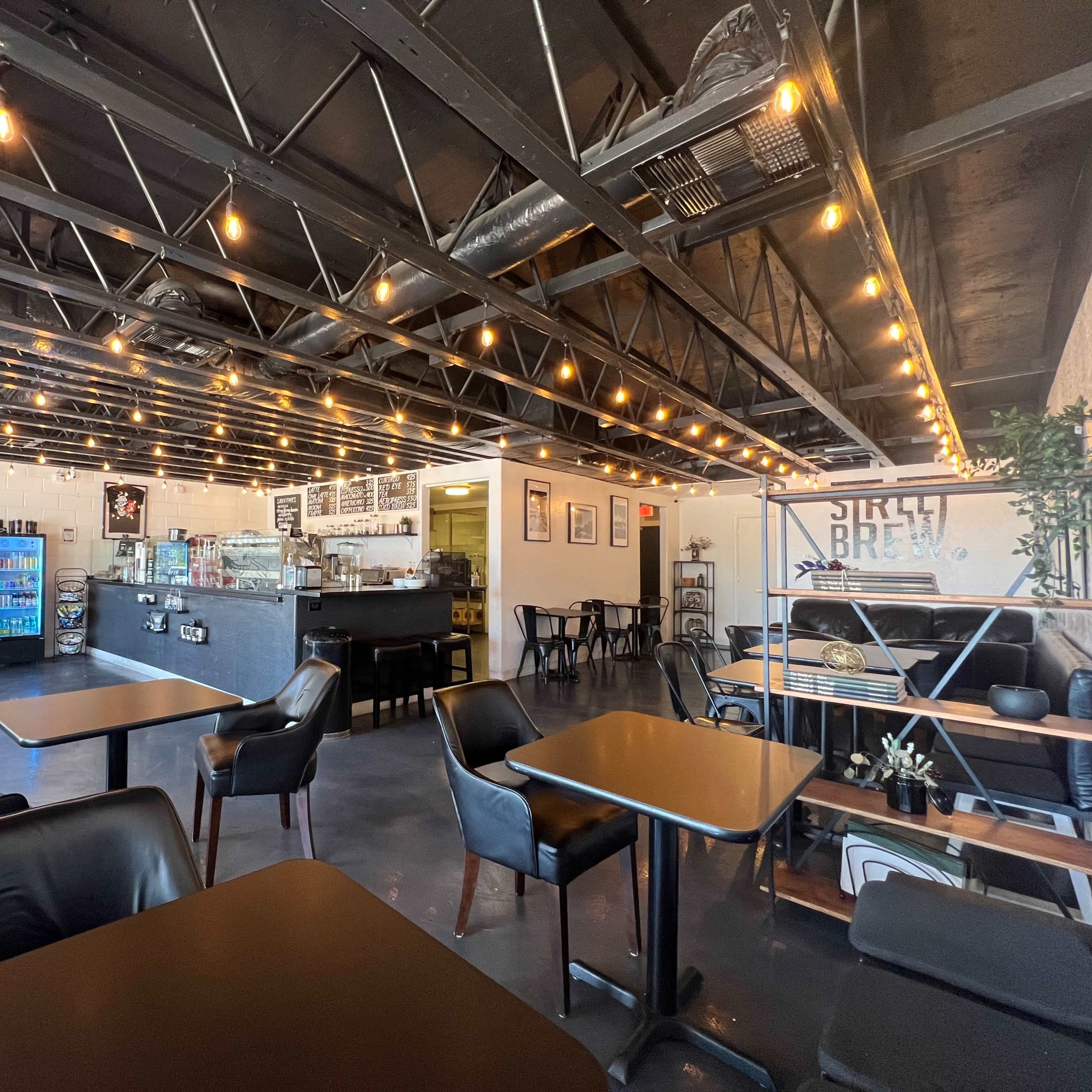 A well-lit cafe interior featuring black tables and chairs, exposed beams, string lights, and a counter with a menu and display cases.