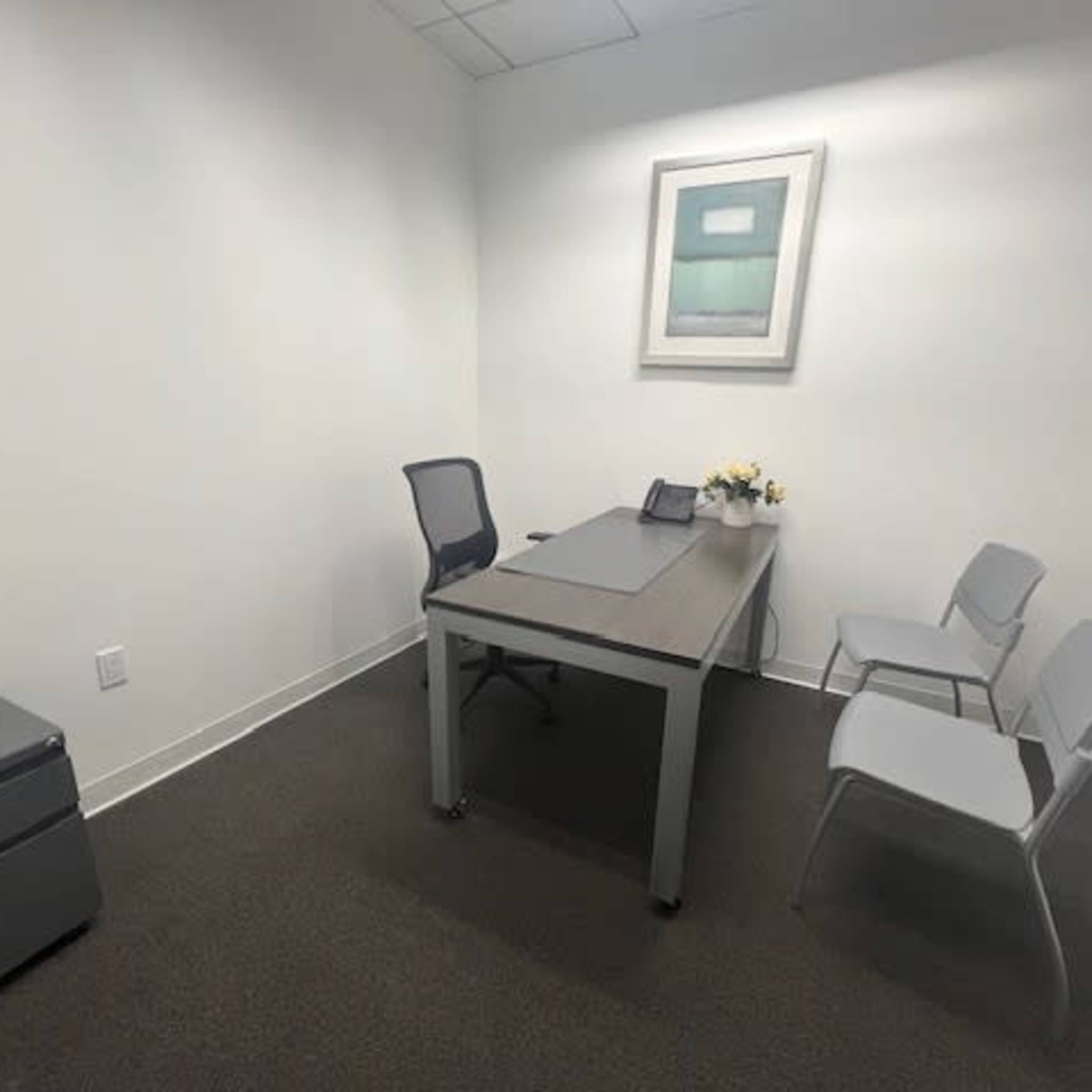 A small office features a desk with a chair, two empty chairs, and a wall art piece above the desk.