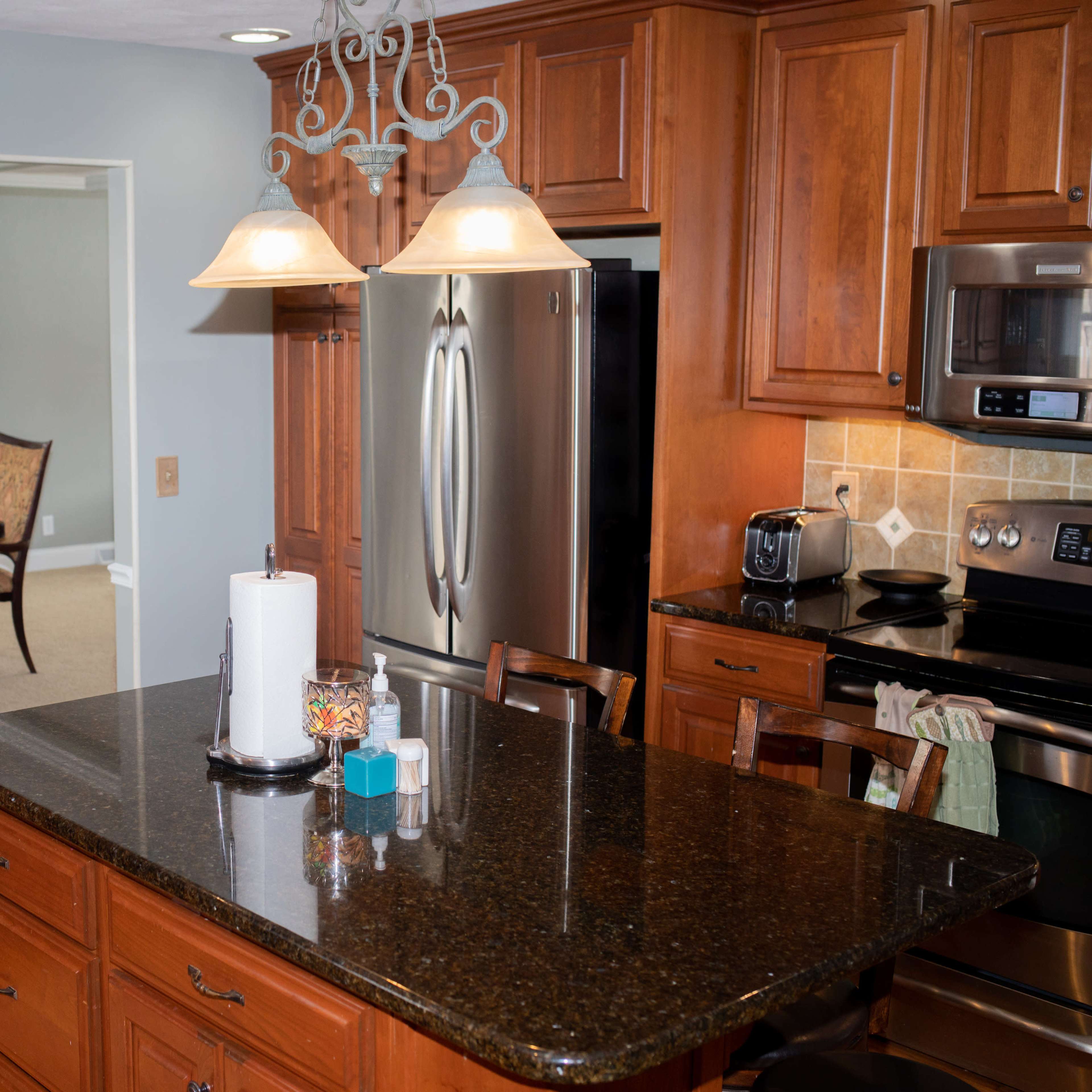 A kitchen with wooden cabinetry, a dark granite countertop, and two pendant lights hanging above, with a dining area visible in the background.
