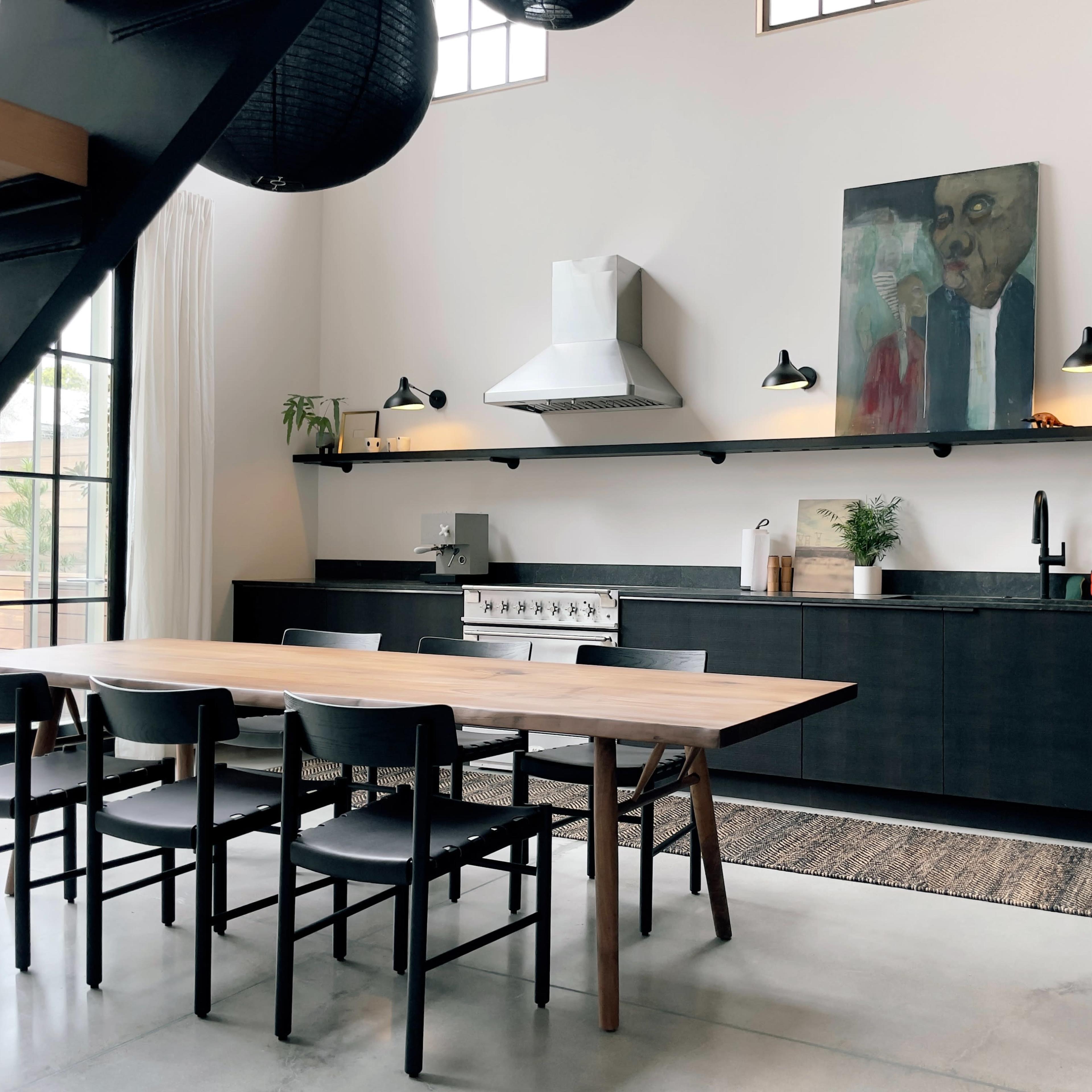 A modern kitchen features a long wooden dining table, black cabinetry, and a large artwork on the wall, with a sleek stove and minimalist decor.