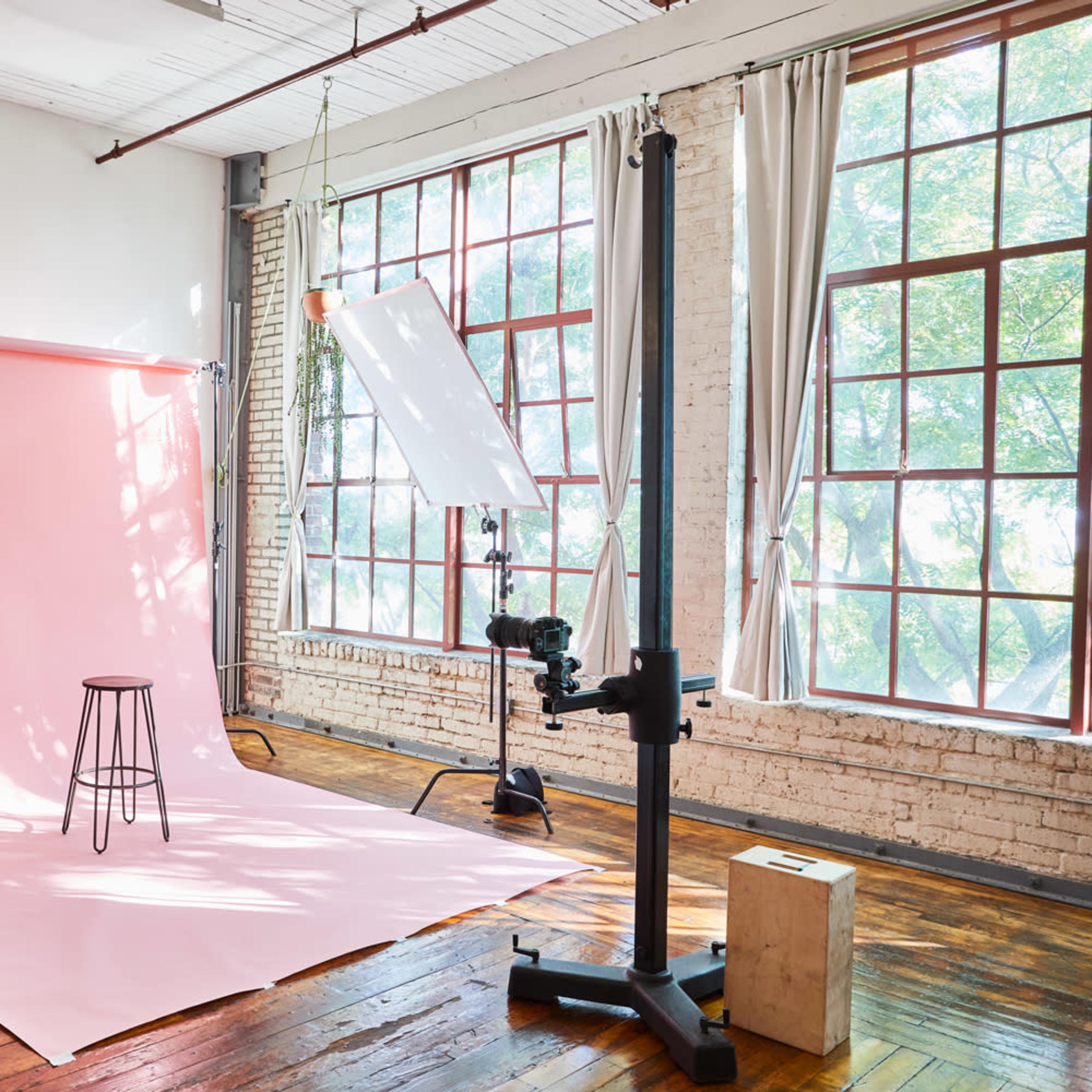 A photography studio setup featuring a pink backdrop, a stool, and various lighting equipment near large windows.