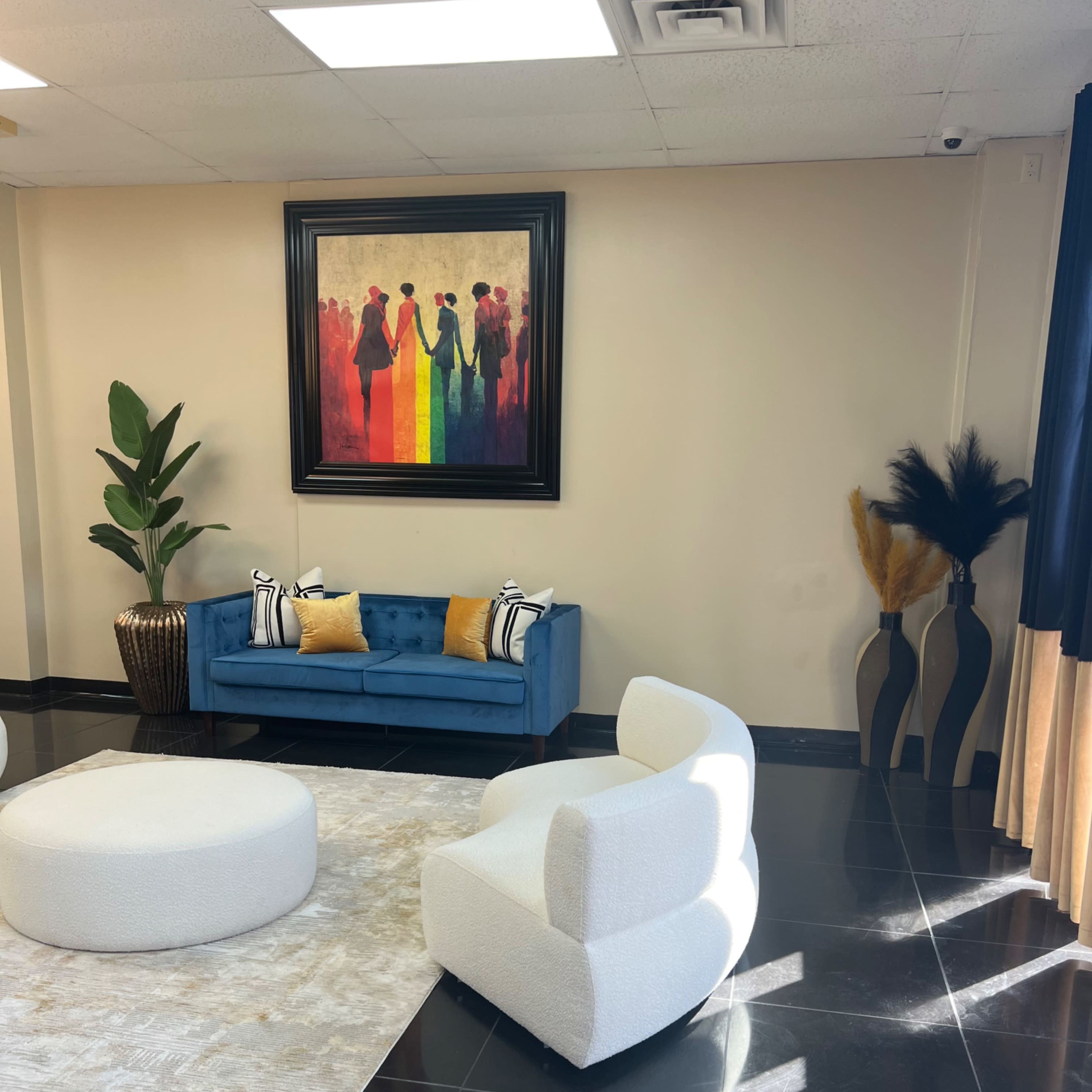 A modern interior space featuring a blue sofa with yellow and black pillows, a round white ottoman, and decorative plants near a large colorful painting on the wall.