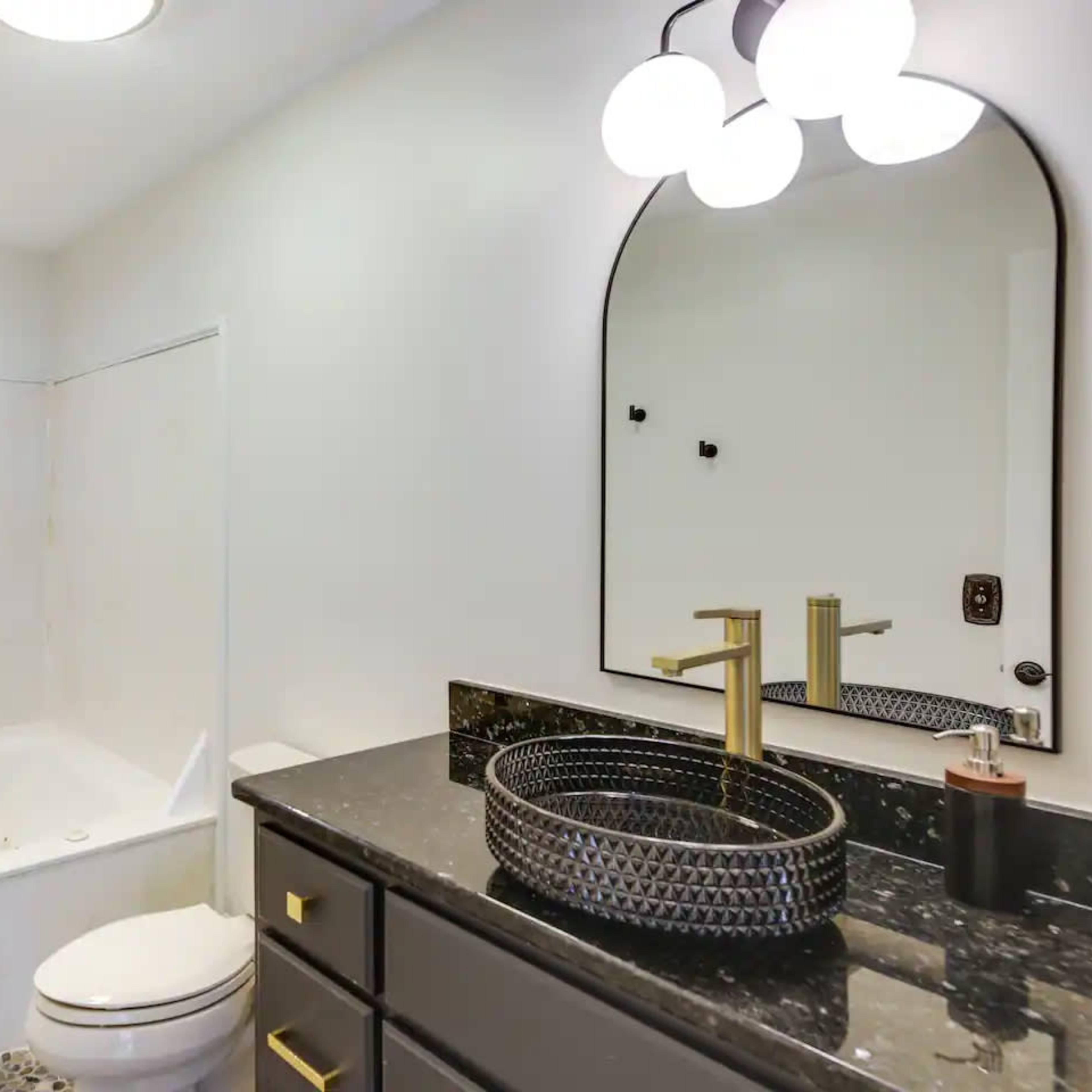 A modern bathroom featuring a bathtub, a wall-mounted mirror, a dark marble countertop, and a stylish faucet beside a round sink.