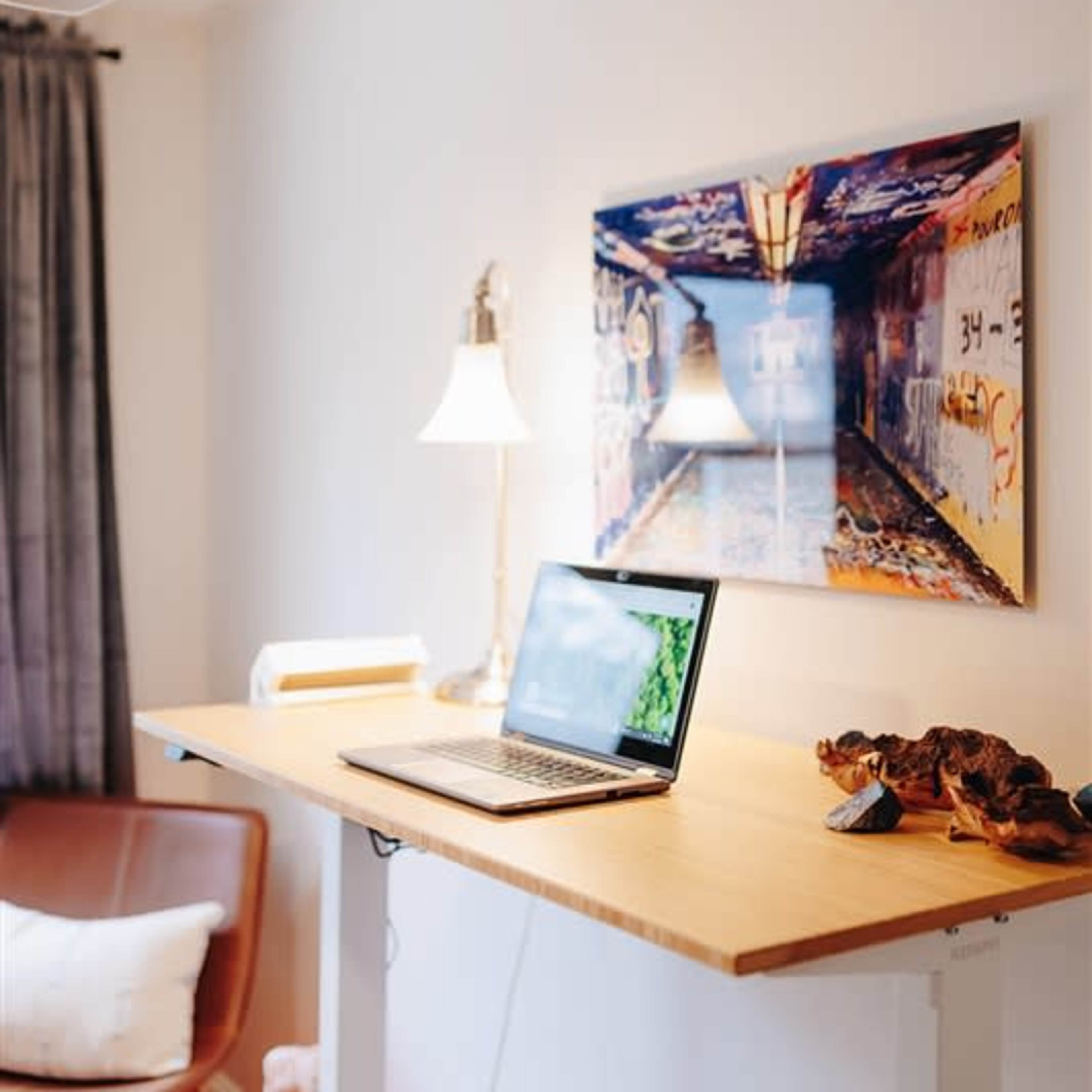 A desk with a laptop is positioned against a wall adorned with a colorful painting, next to a lamp and a chair in a well-lit room.
