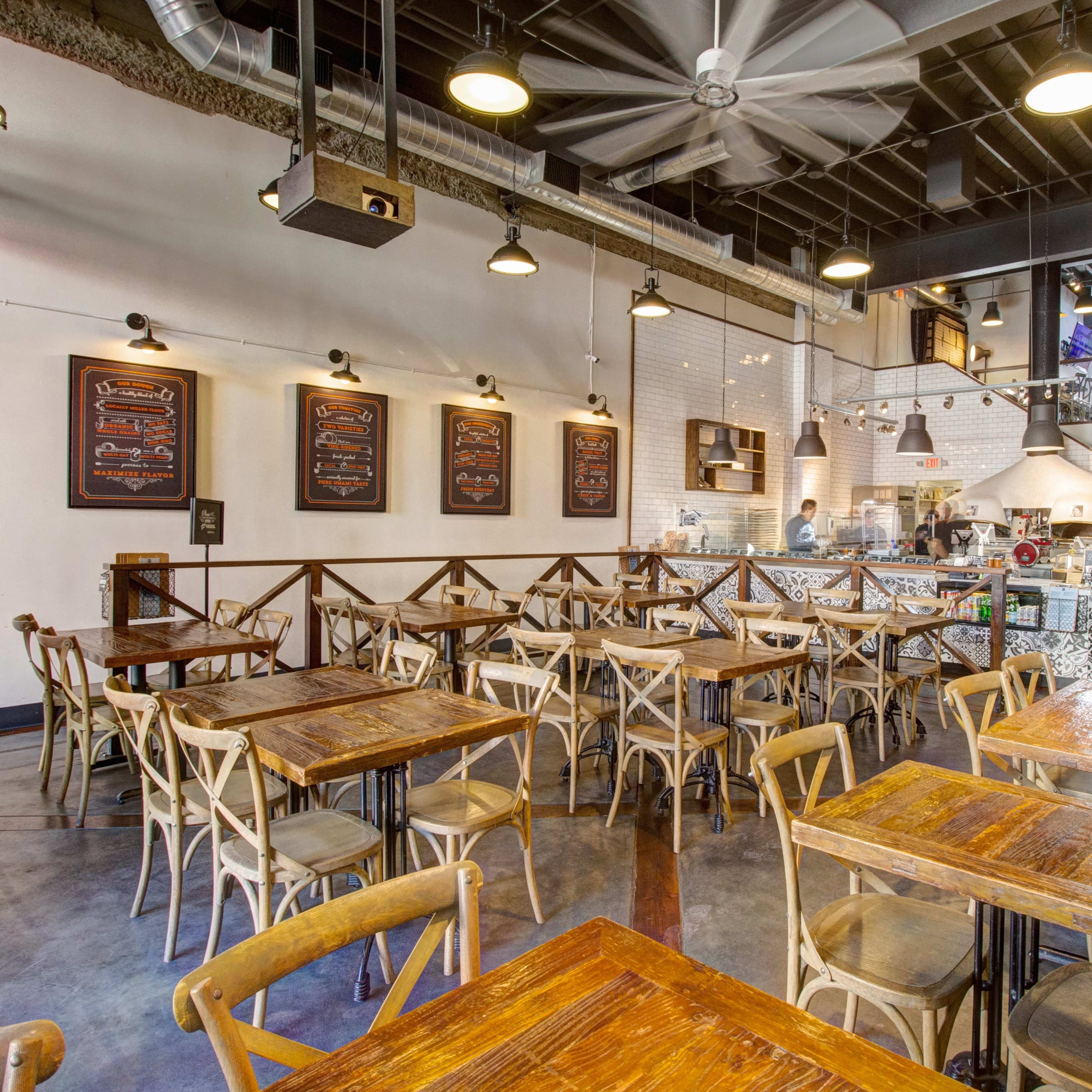 A spacious restaurant interior featuring wooden tables and chairs, with a visible kitchen area in the background and menus displayed on the walls.
