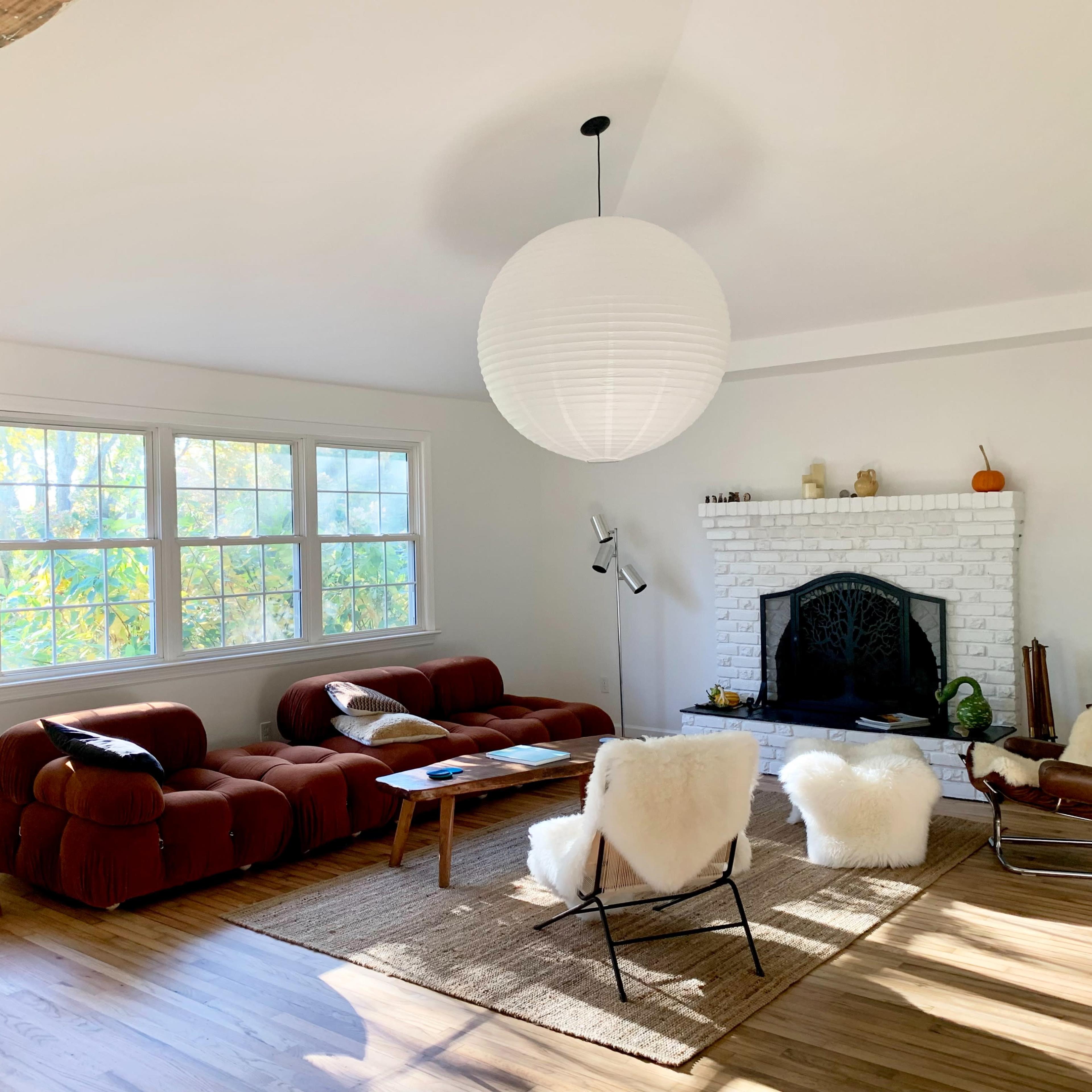 A modern living room features a brown sectional couch, white fireplace with a mantel, and a large pendant light, with sunlight streaming through the windows.