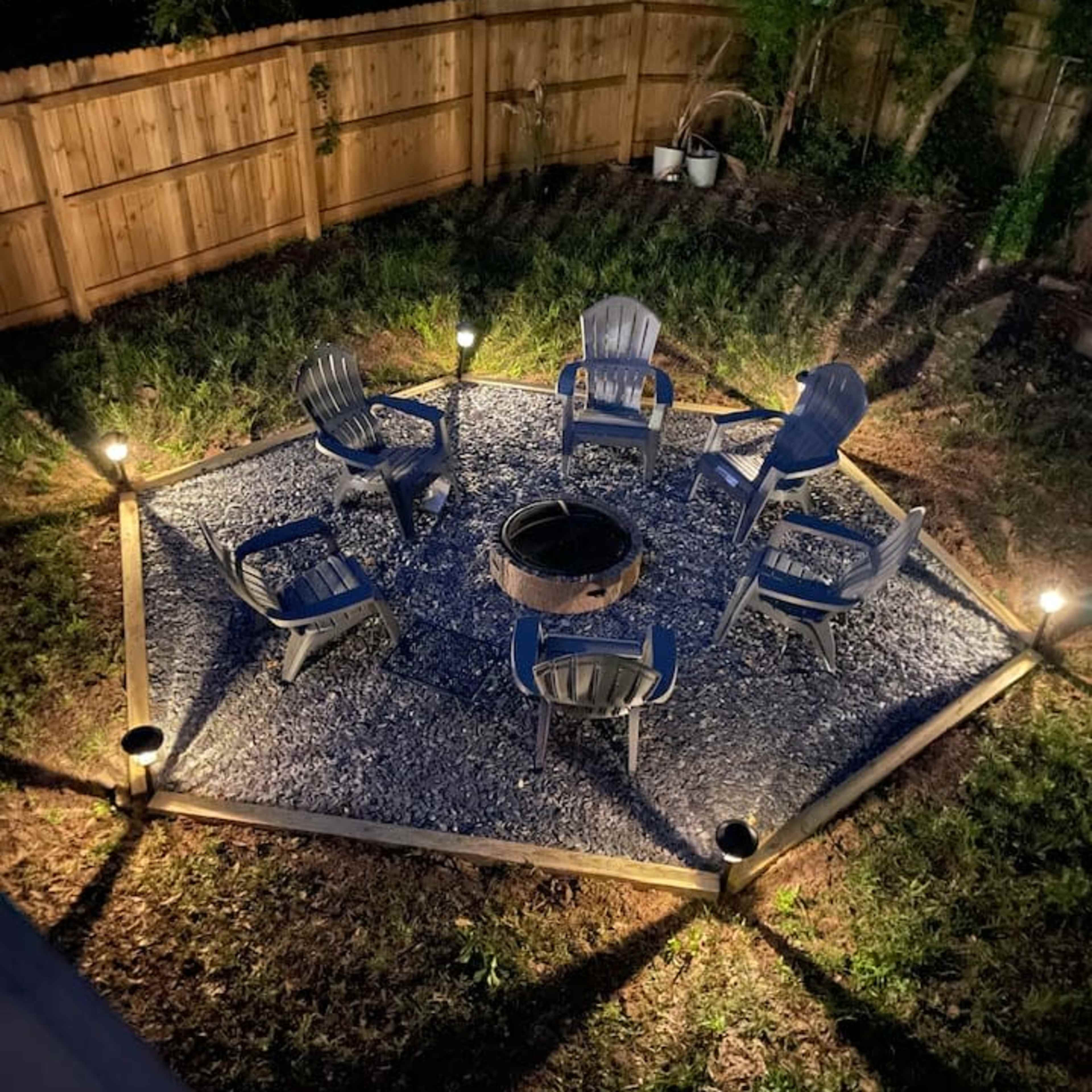 A circular stone fire pit area is surrounded by six gray outdoor chairs, illuminated by lights along the perimeter.
