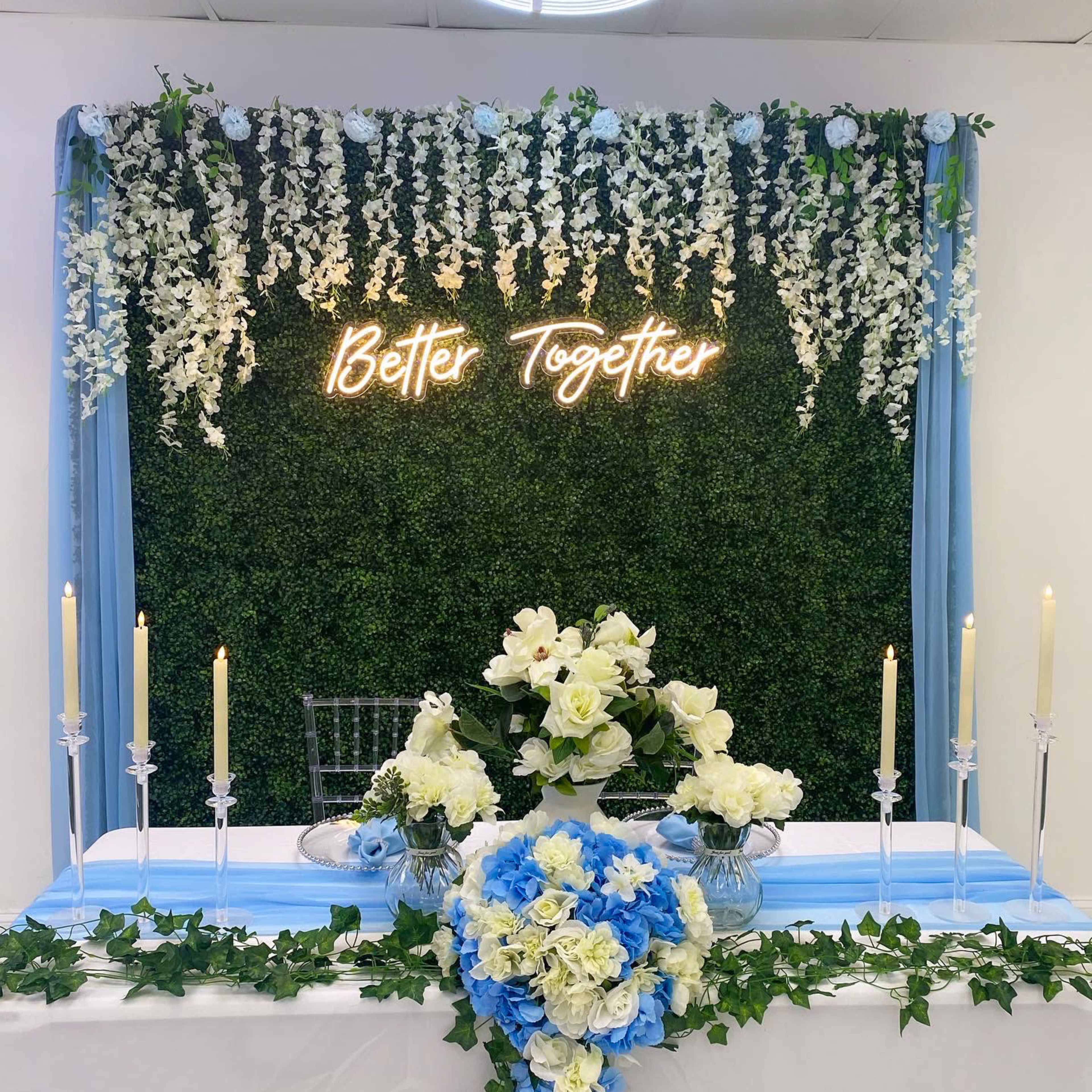 A decorated wedding table is set against a floral backdrop with the phrase "Better Together" illuminated in neon lights.