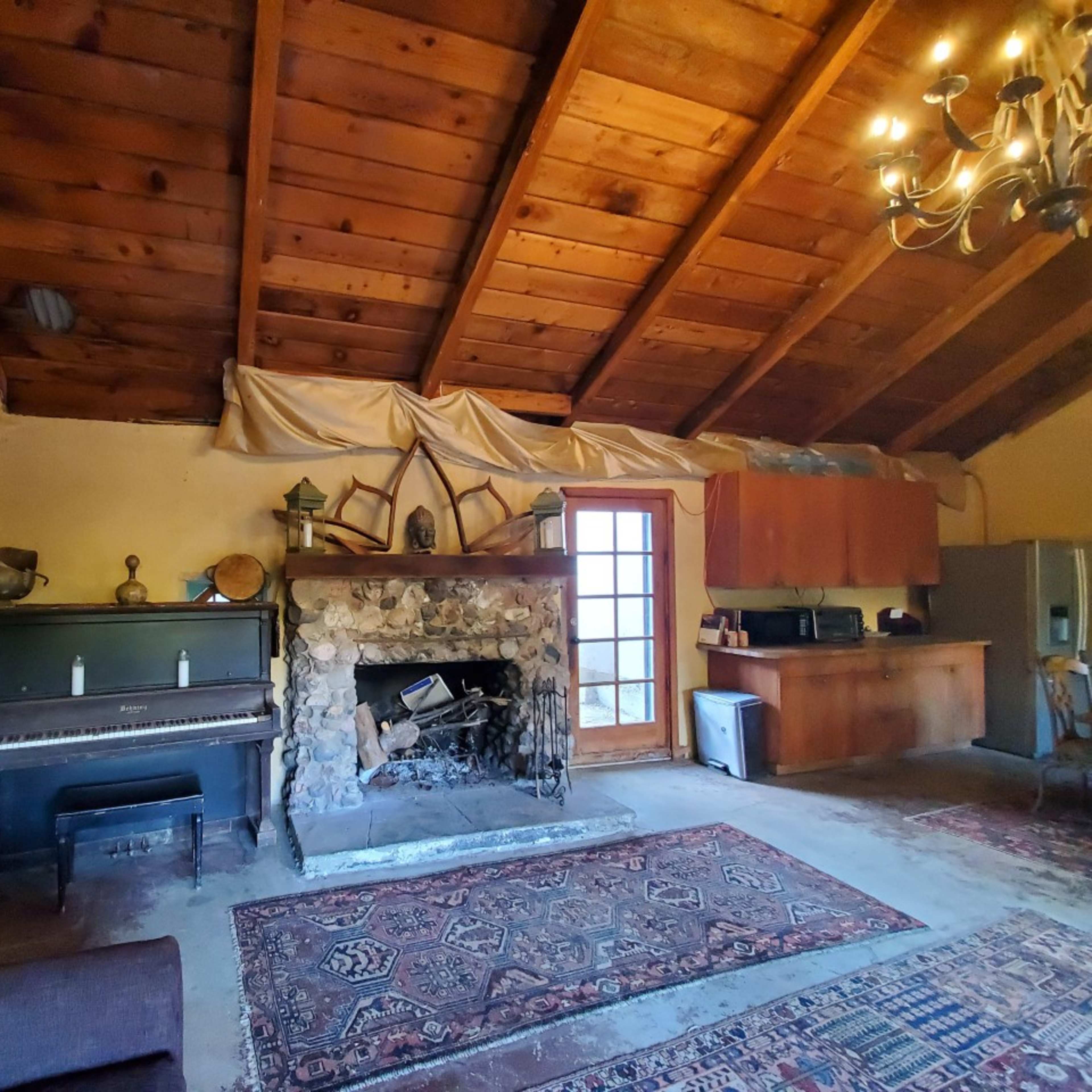 The room features a wooden ceiling with exposed beams, a stone fireplace, a piano, and a small kitchen area along one side.