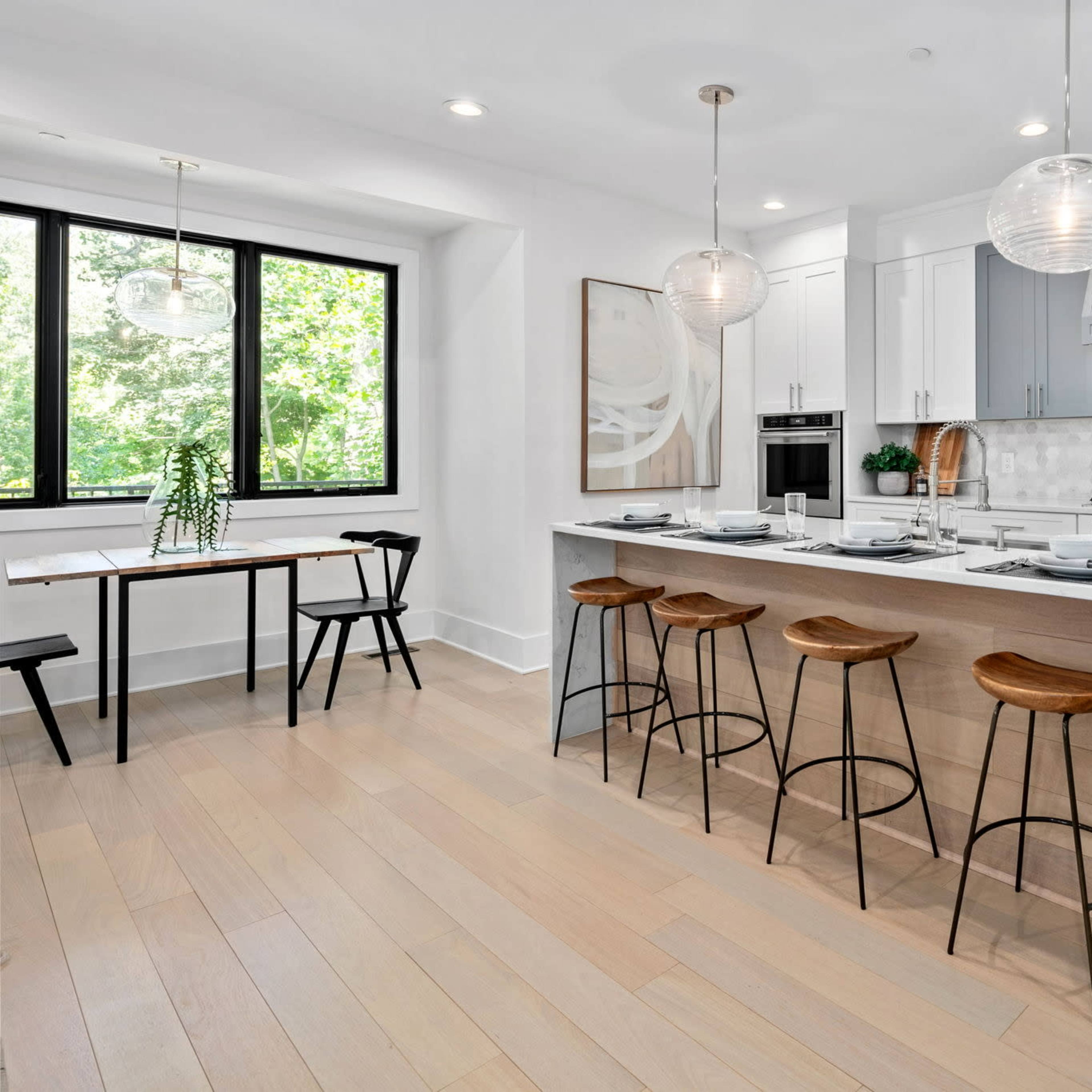 A modern kitchen with a large island, four bar stools, and a dining table near a window overlooking greenery.