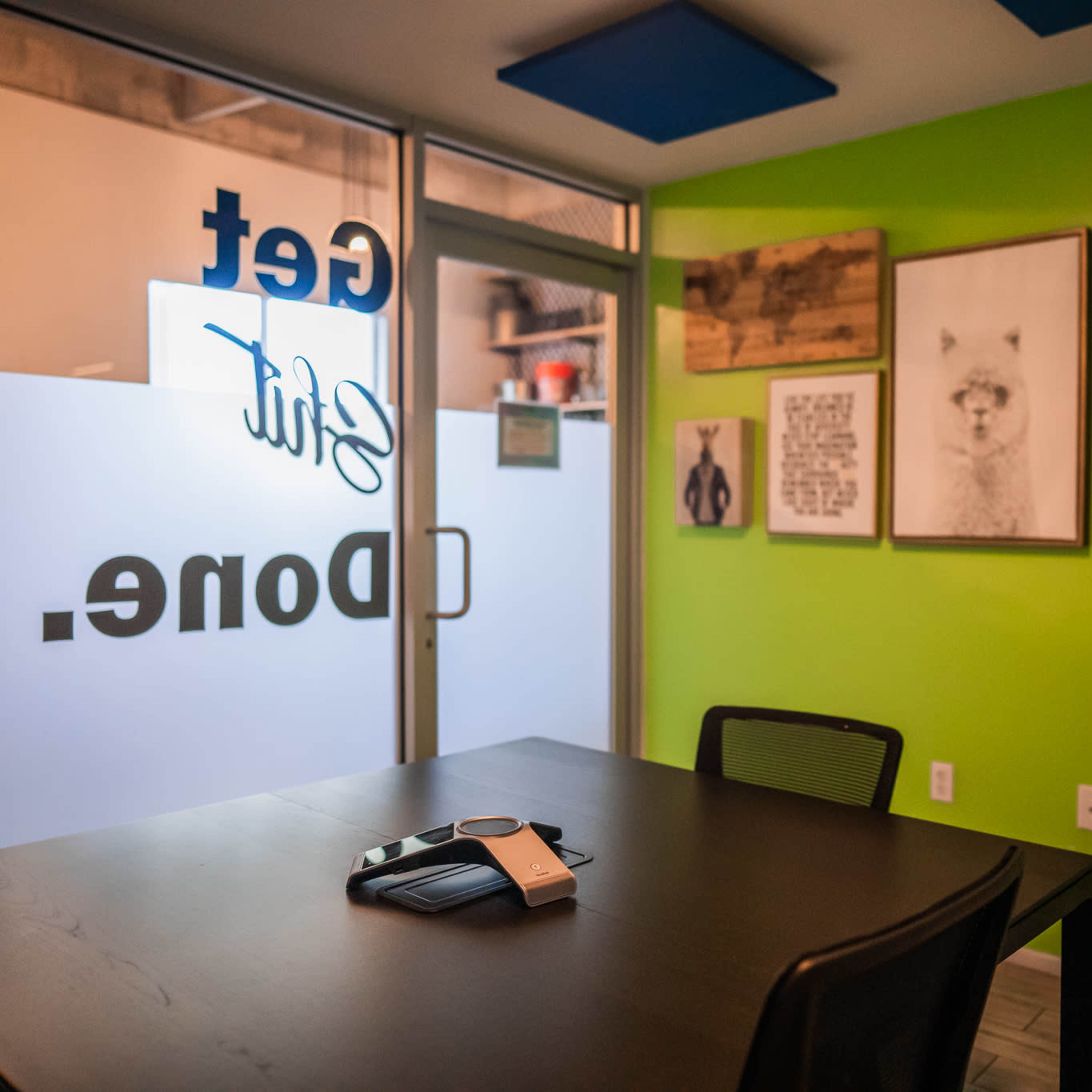 A meeting room features a black table with a phone and several framed artworks on bright green walls.
