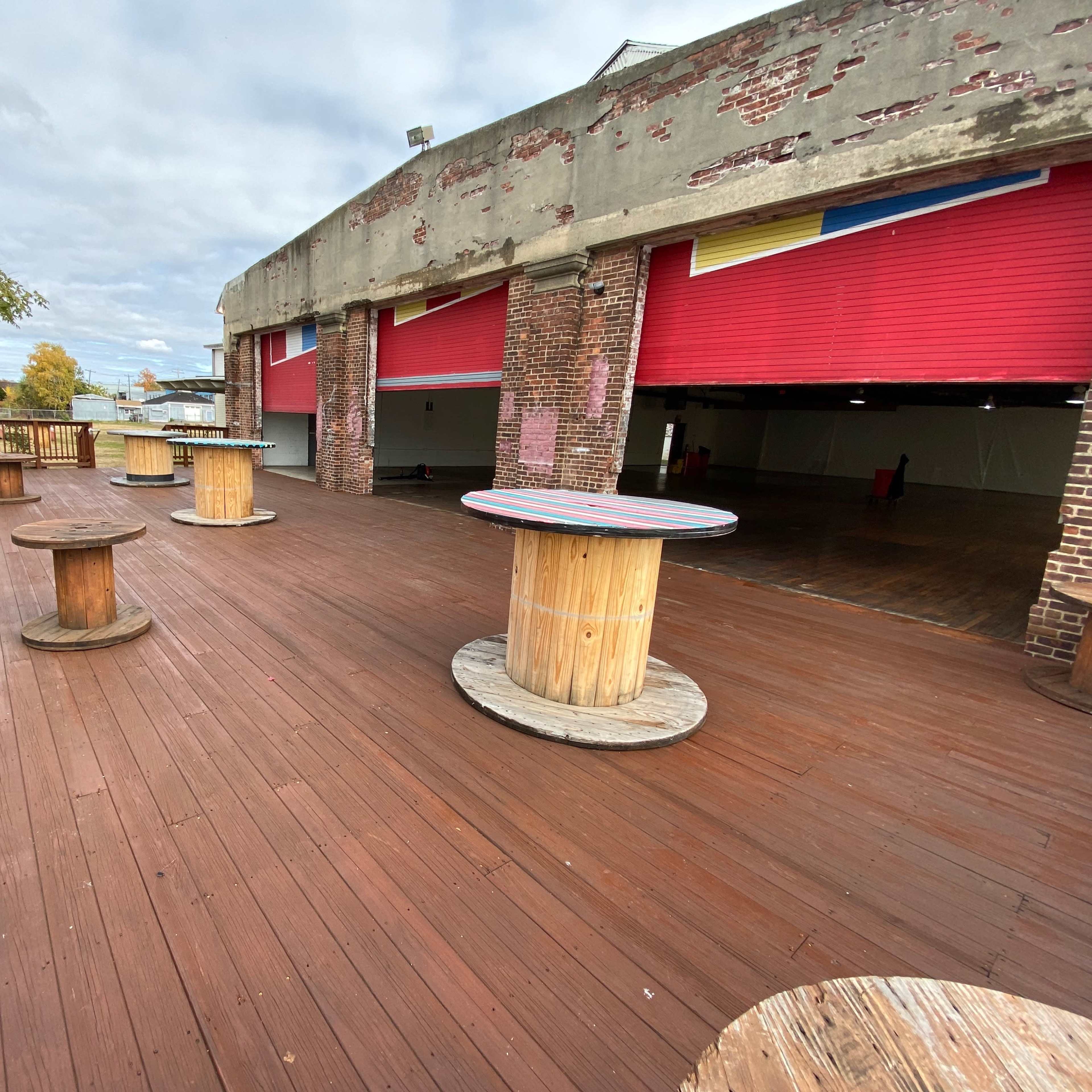 A wooden deck area with several round tables made from large cable spools, set against a backdrop of brick walls and red garage doors.