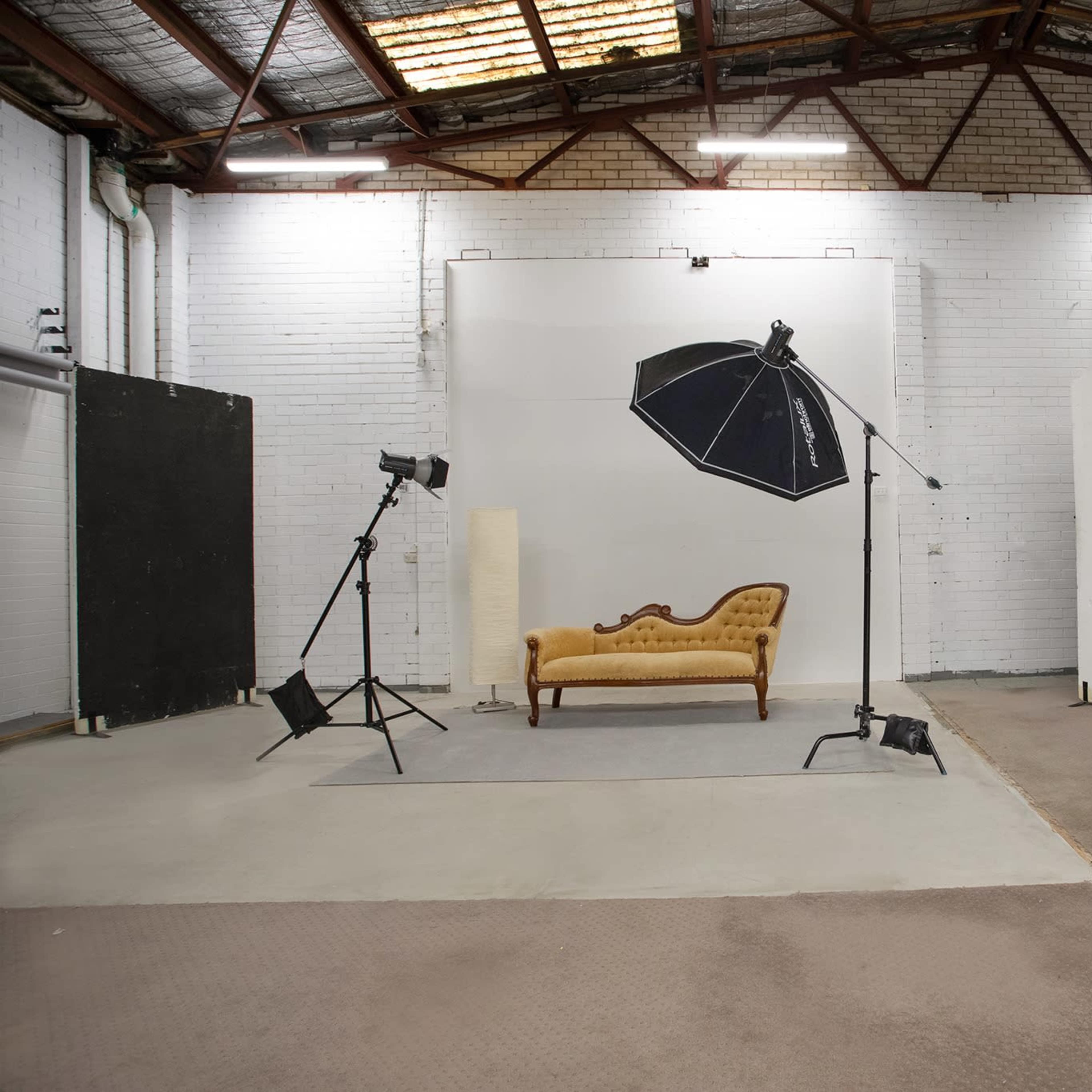 A photo studio featuring a vintage-style couch, a large umbrella light, and various backdrops in a spacious, industrial setting.