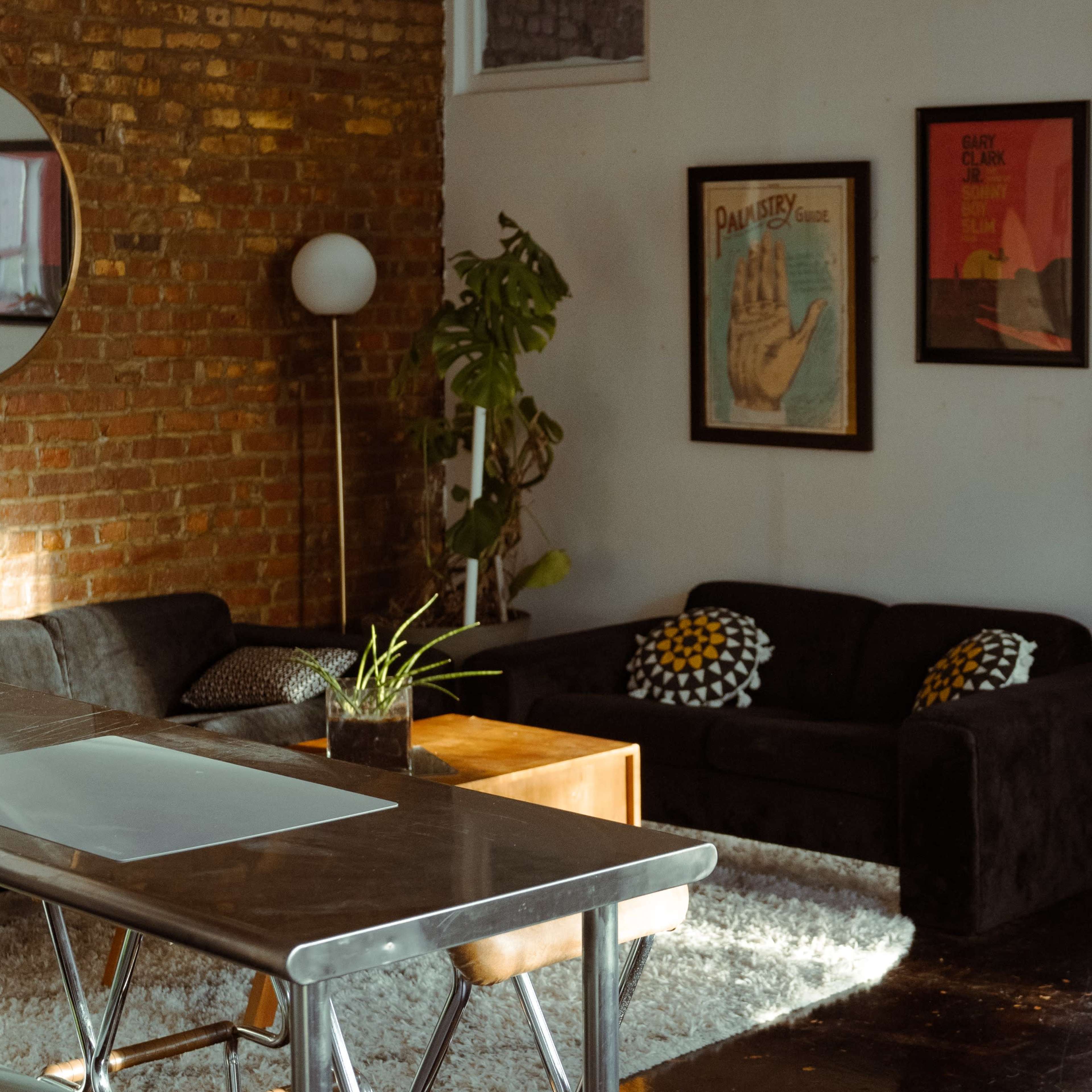 A modern living space features a kitchen area with a stainless steel table and microwave, adjacent to a cozy seating area with dark sofas and framed artwork on the exposed brick wall.