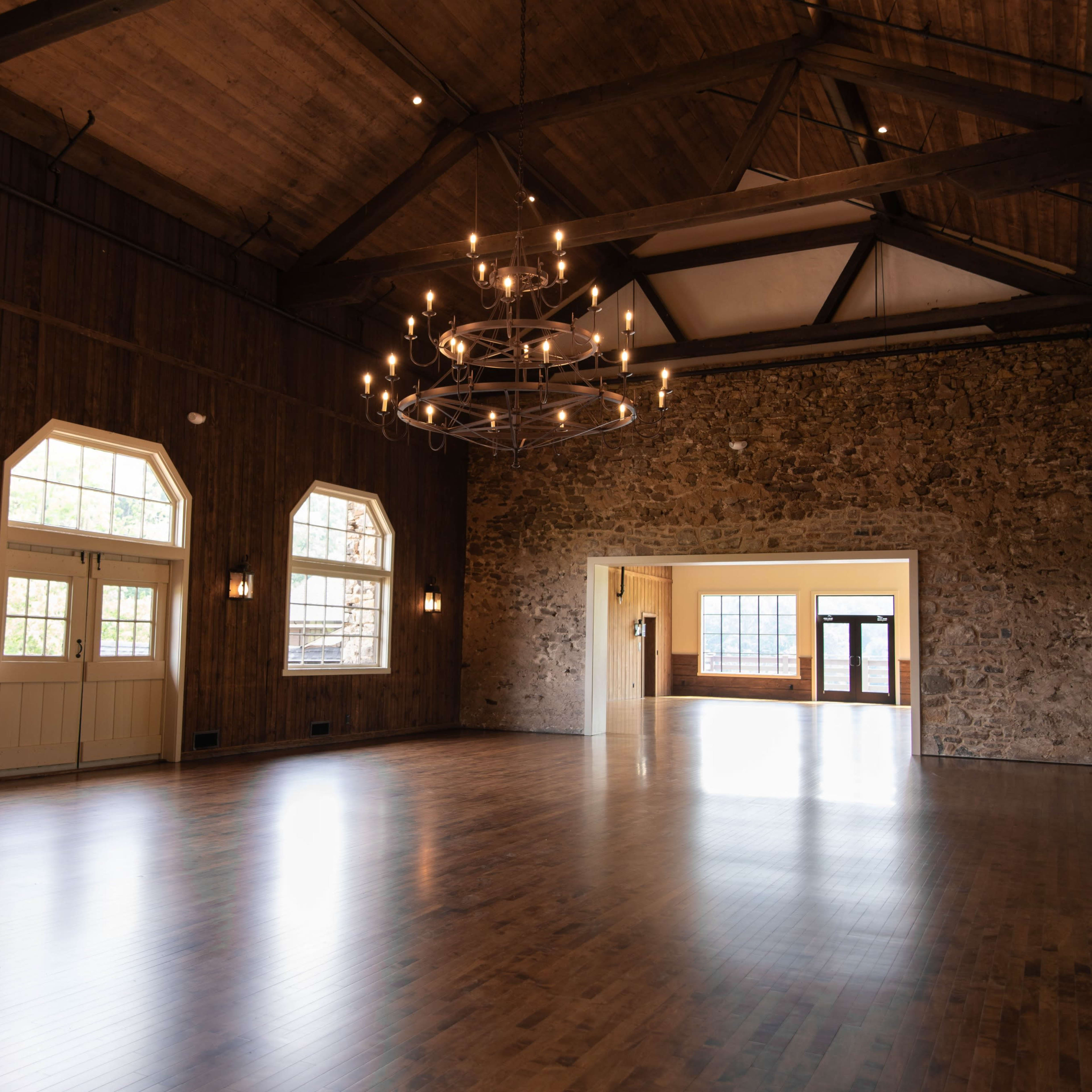 A spacious indoor hall with wooden beams, large windows, and a stone accent wall, featuring polished wooden floors and a modern chandelier.