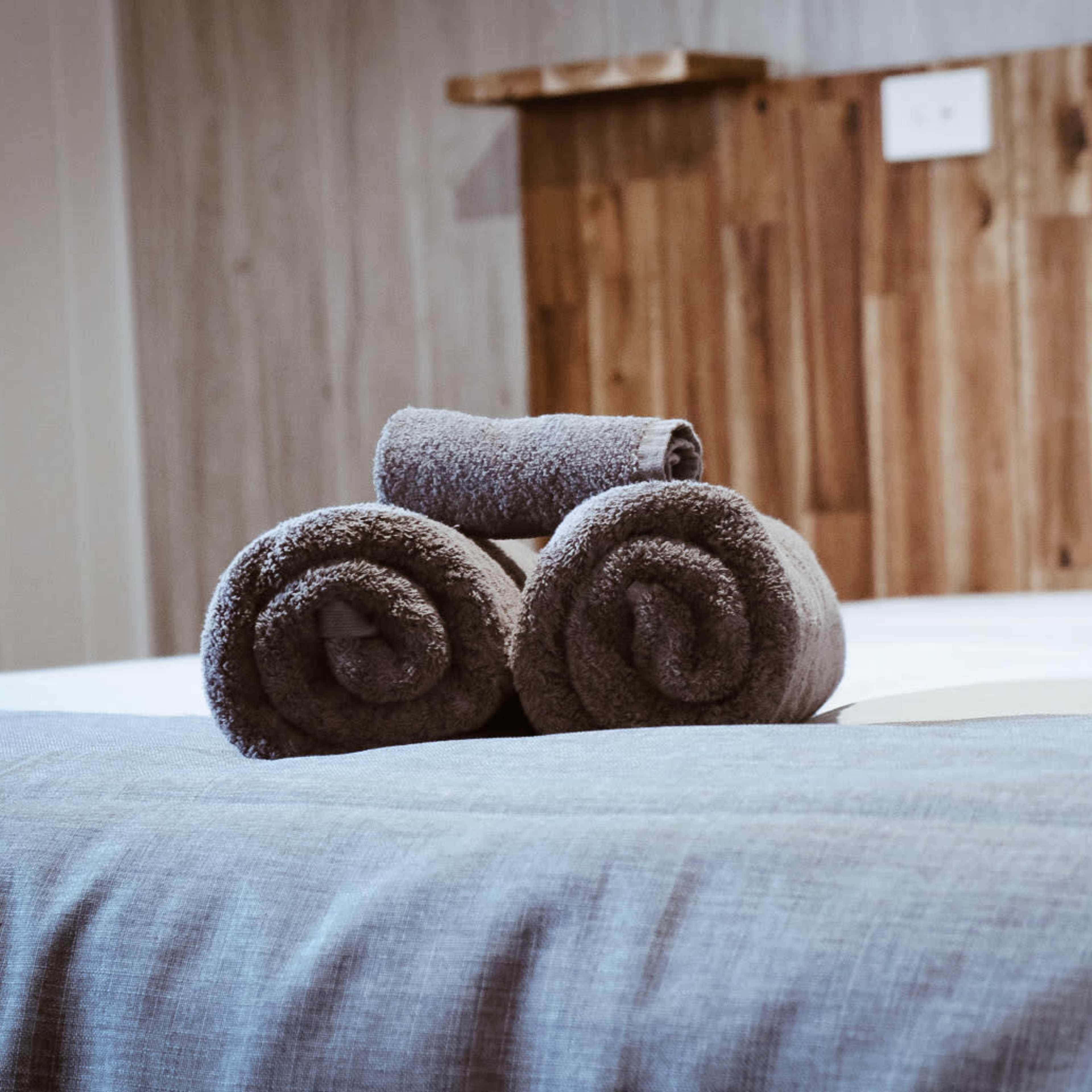 Three neatly rolled gray towels are placed on a neatly made bed with a wooden headboard.