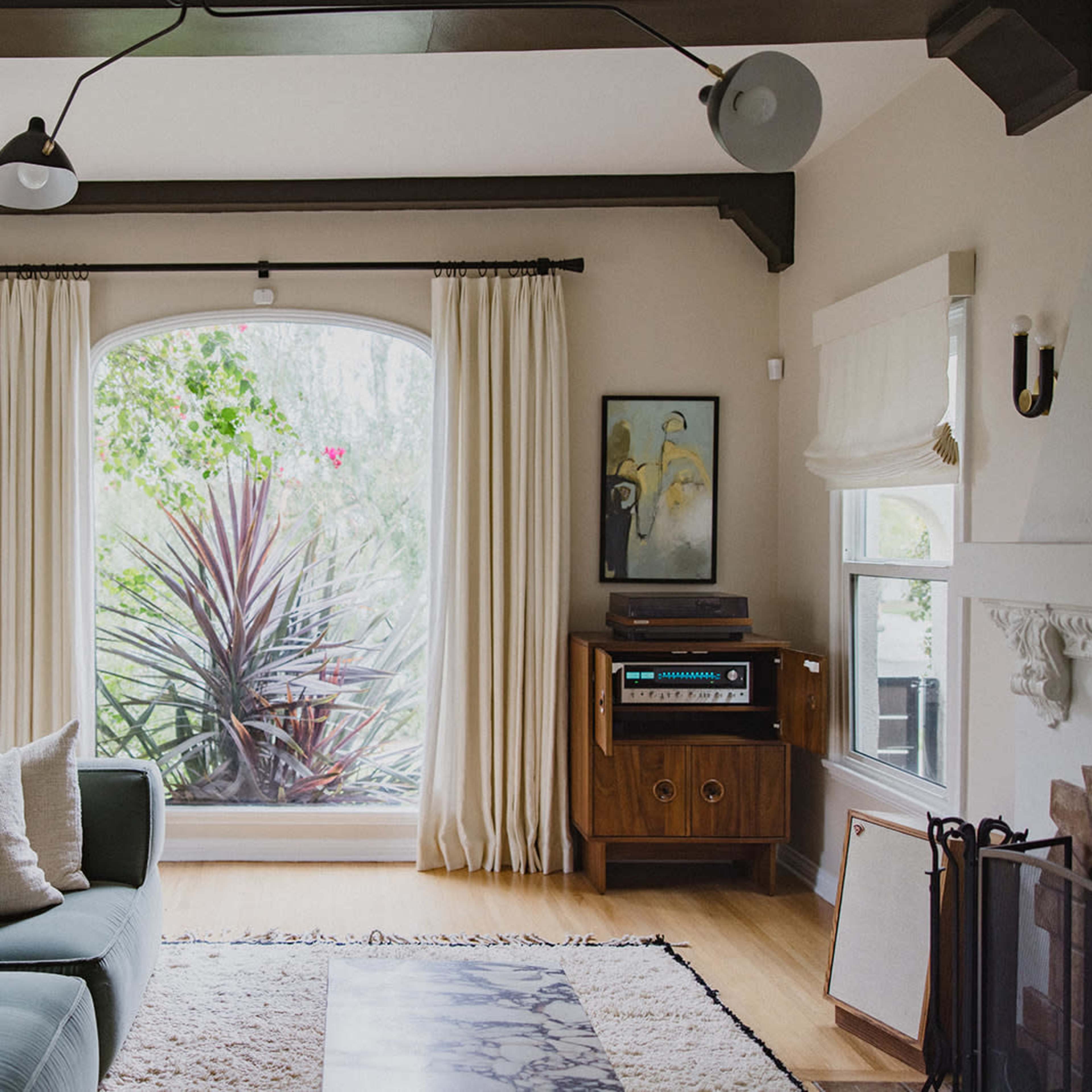 A light-filled living room features a green sectional sofa, large windows with curtains, and a vintage stereo unit beside a wooden cabinet.