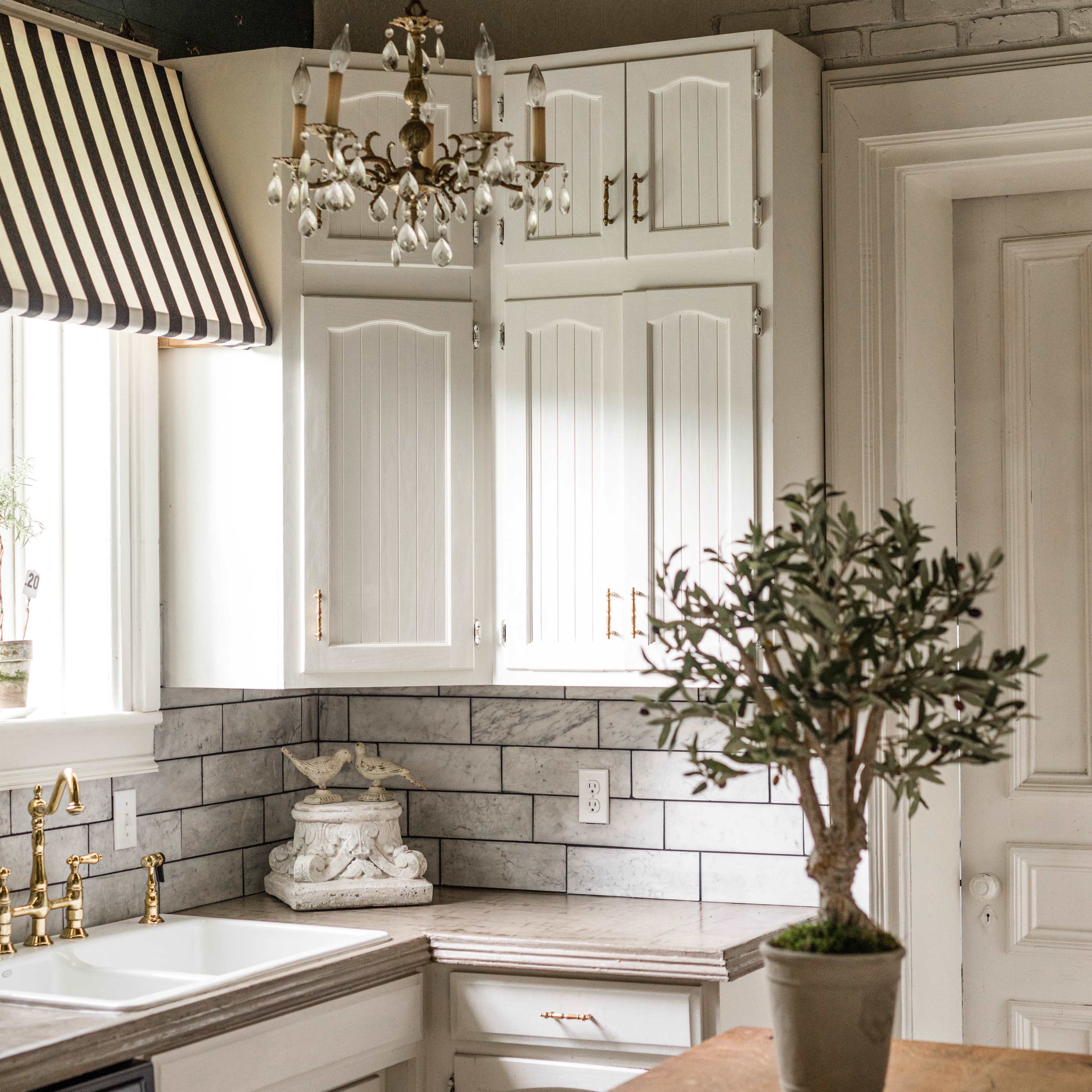 A well-lit kitchen featuring white cabinetry, a farmhouse sink, and a wooden table, with a potted plant and decorative elements in the background.