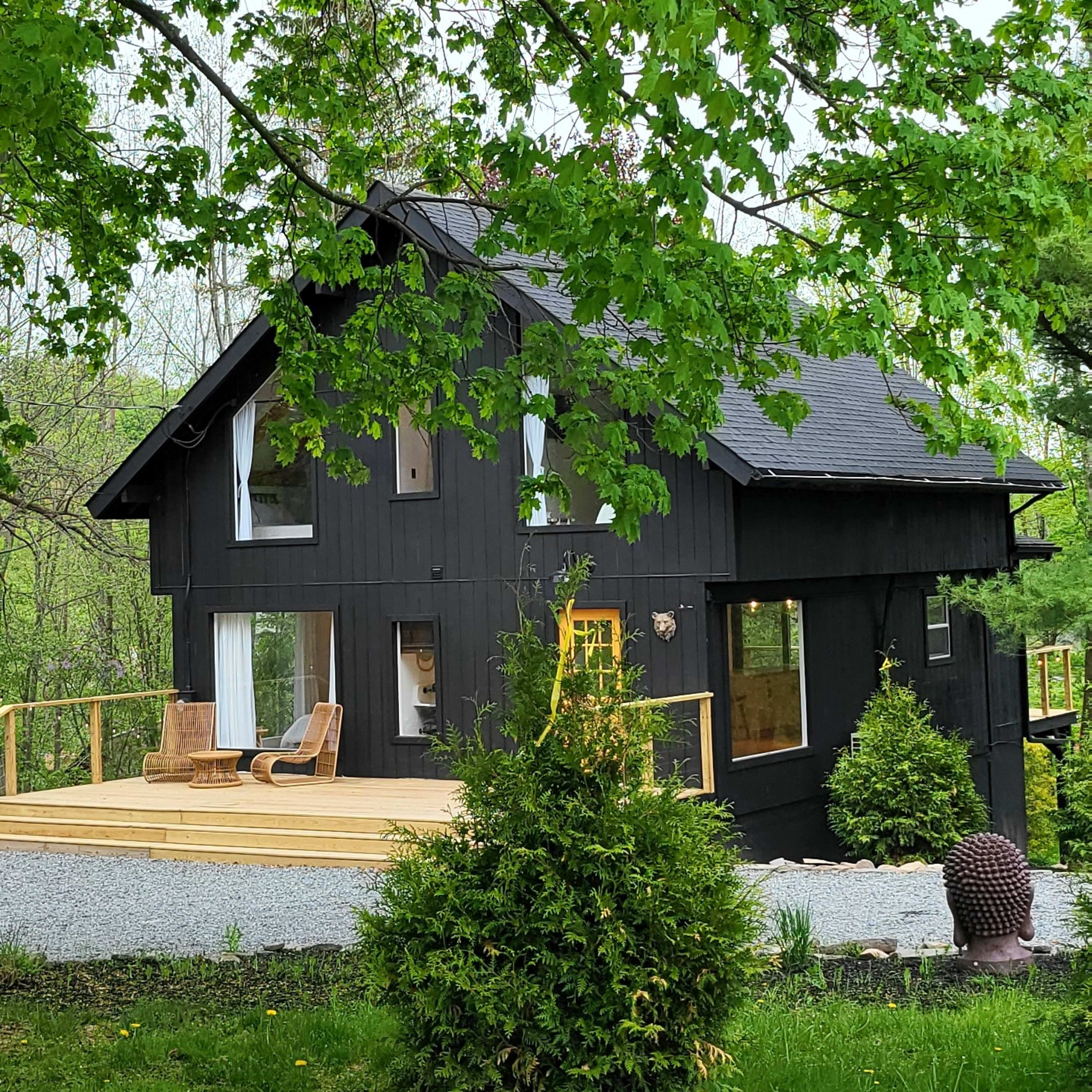 A dark-colored house surrounded by greenery, set back from the road with a wooden deck and large windows.