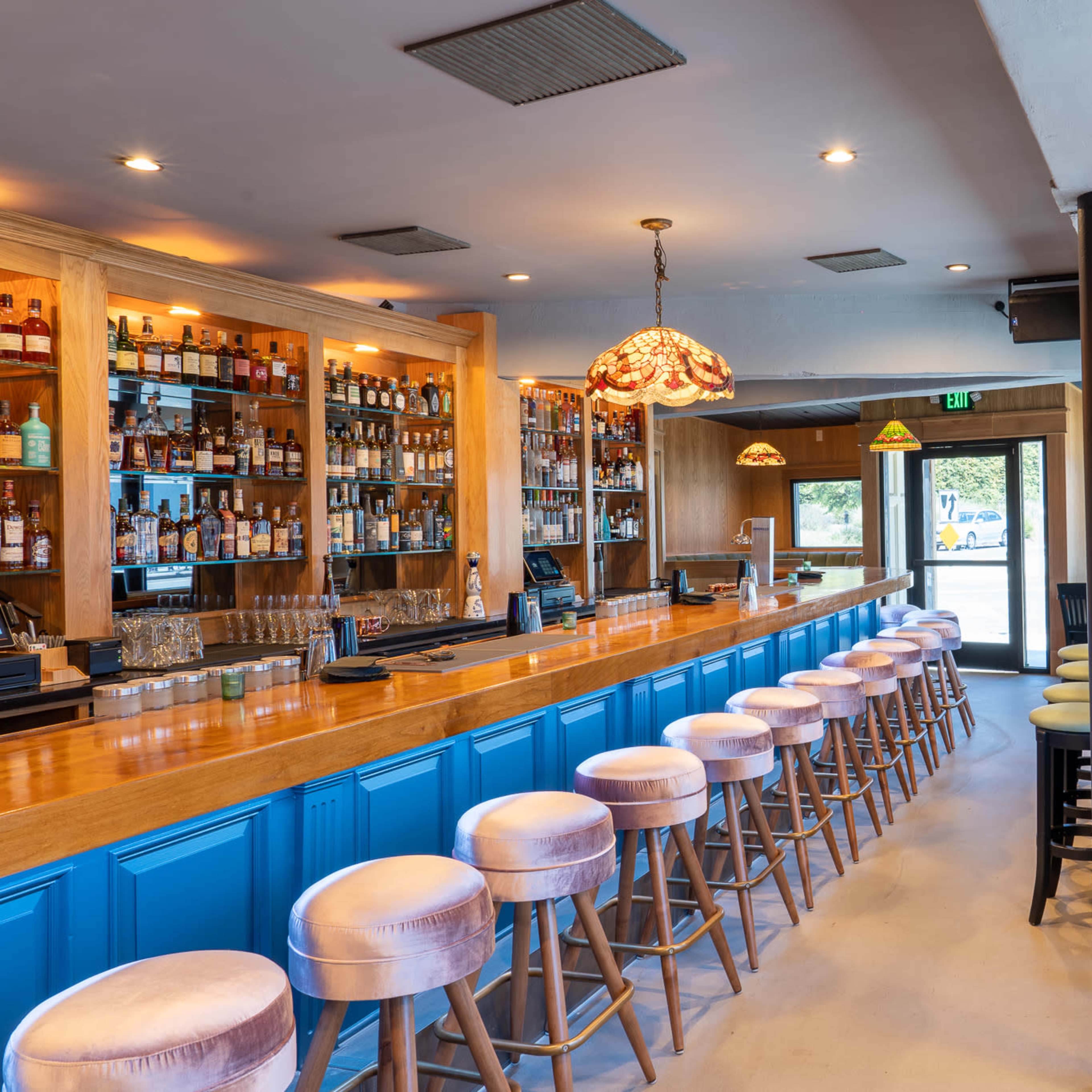 A spacious bar area with a wooden counter, high stools, and shelves lined with various bottles, illuminated by decorative pendant lights.