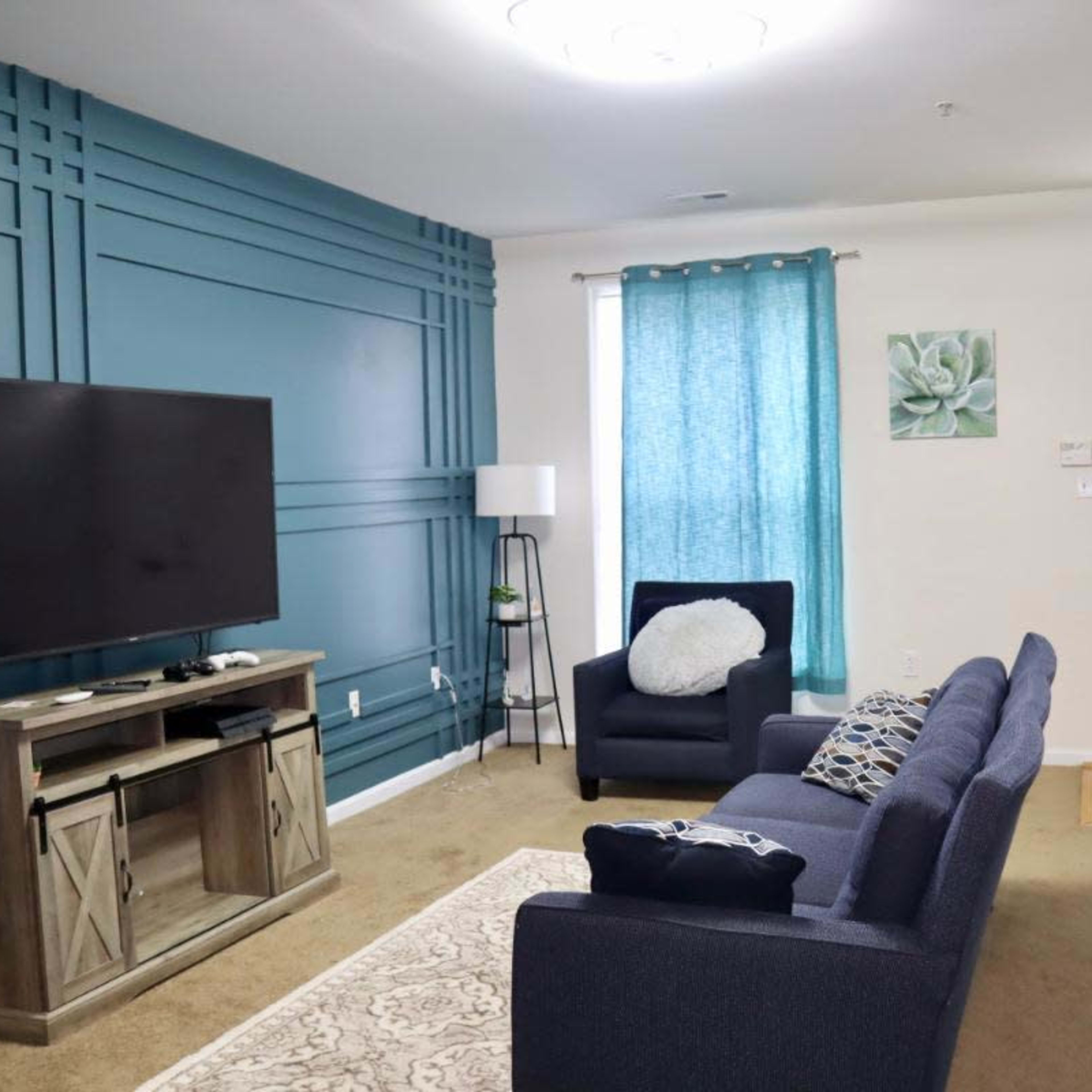 A living room with a blue accent wall, a dark sofa facing a TV, a light-colored area rug, and a window with teal curtains.