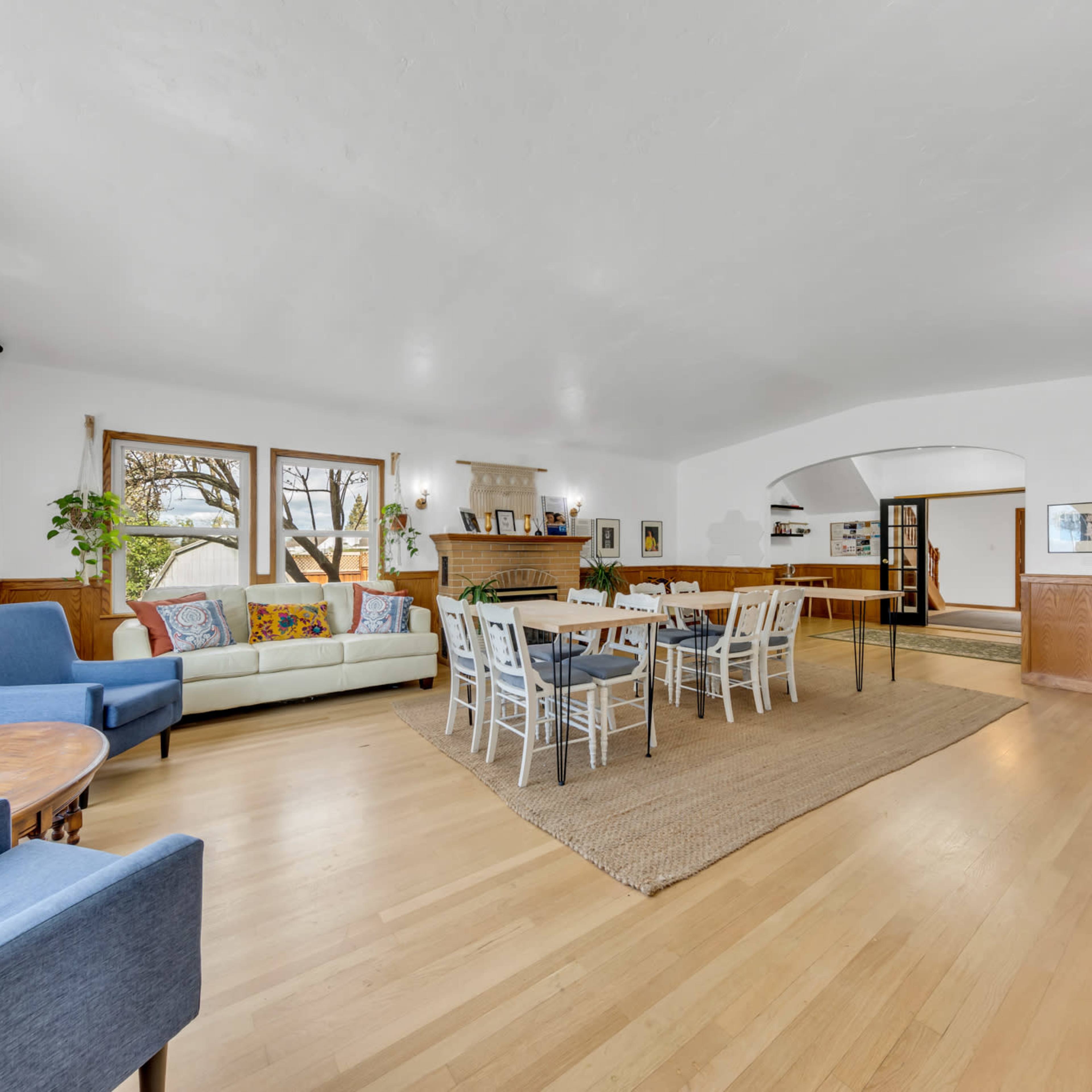 The spacious living area features a light-colored wooden floor, large windows, and a mix of seating arrangements, including a sofa and dining tables.