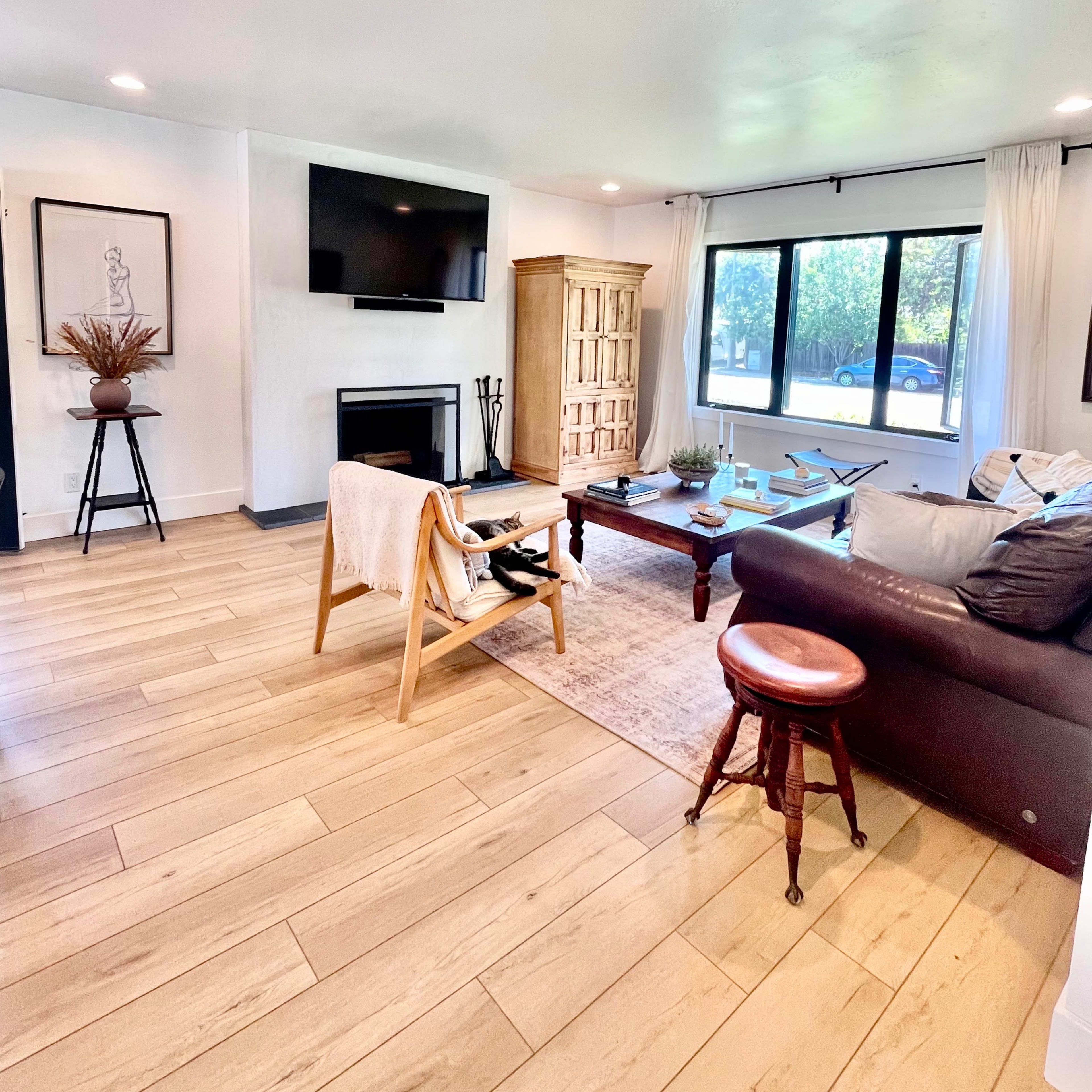 A bright living room with a flat-screen TV mounted on the wall, a wooden cabinet, and a seating area featuring a leather sofa and a wooden table.