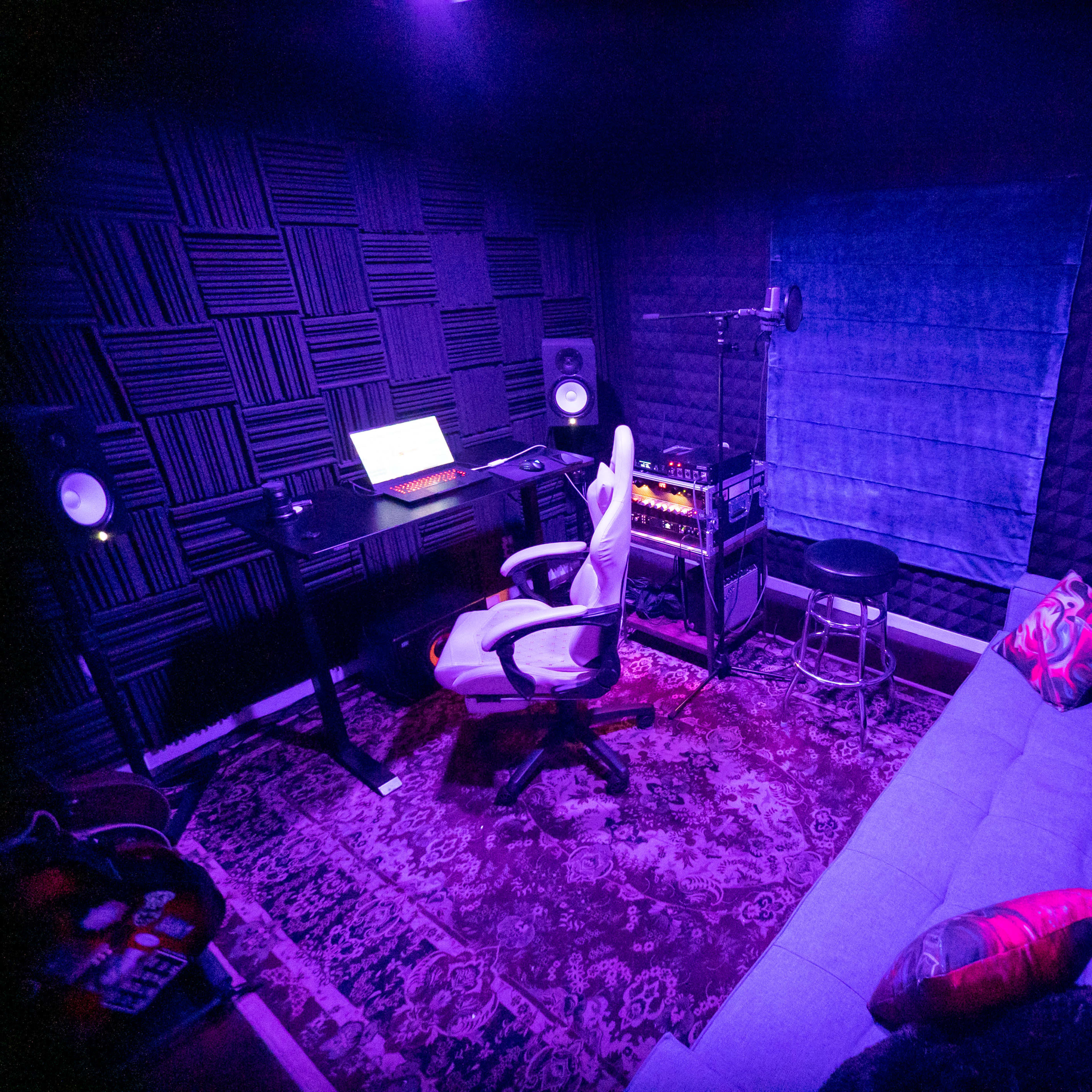 A music production studio features a desk with a laptop, speakers, and a cozy couch, all illuminated by purple lighting.