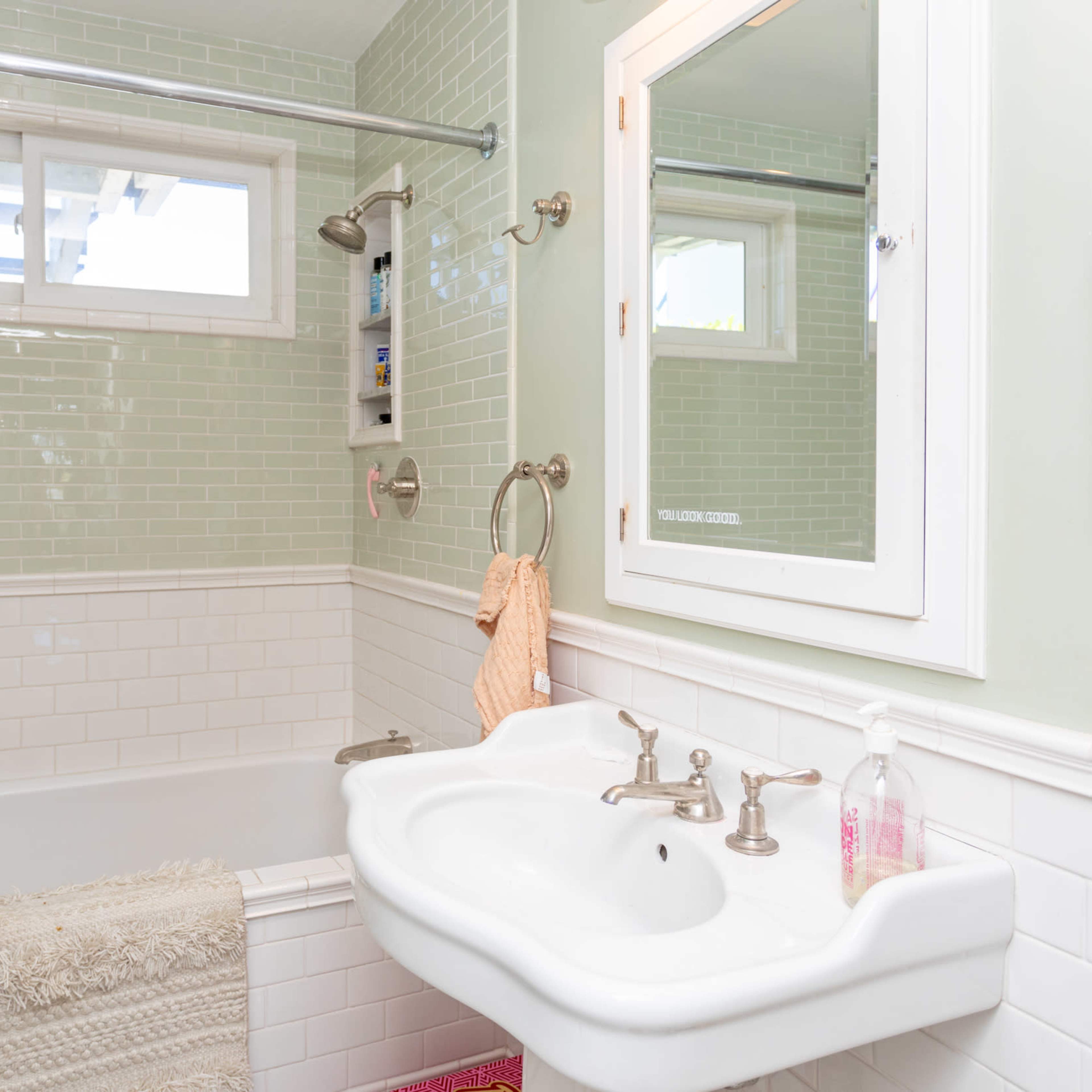 A bathroom featuring a white bathtub, a wall-mounted mirror, and light green tiled walls with a window above the tub.