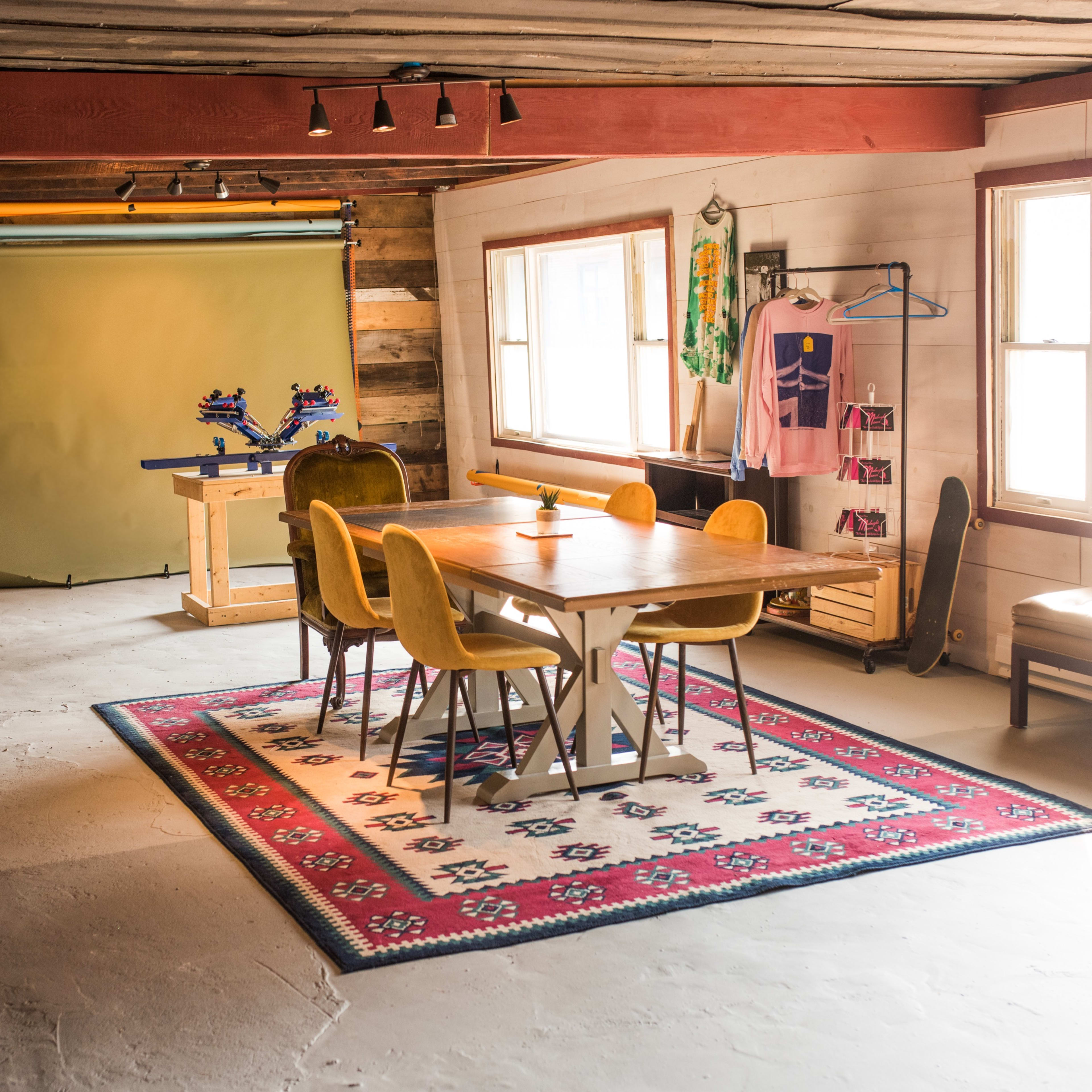 A spacious room with a wooden table surrounded by yellow chairs, a light-colored rug, and a backdrop for photography in the background.