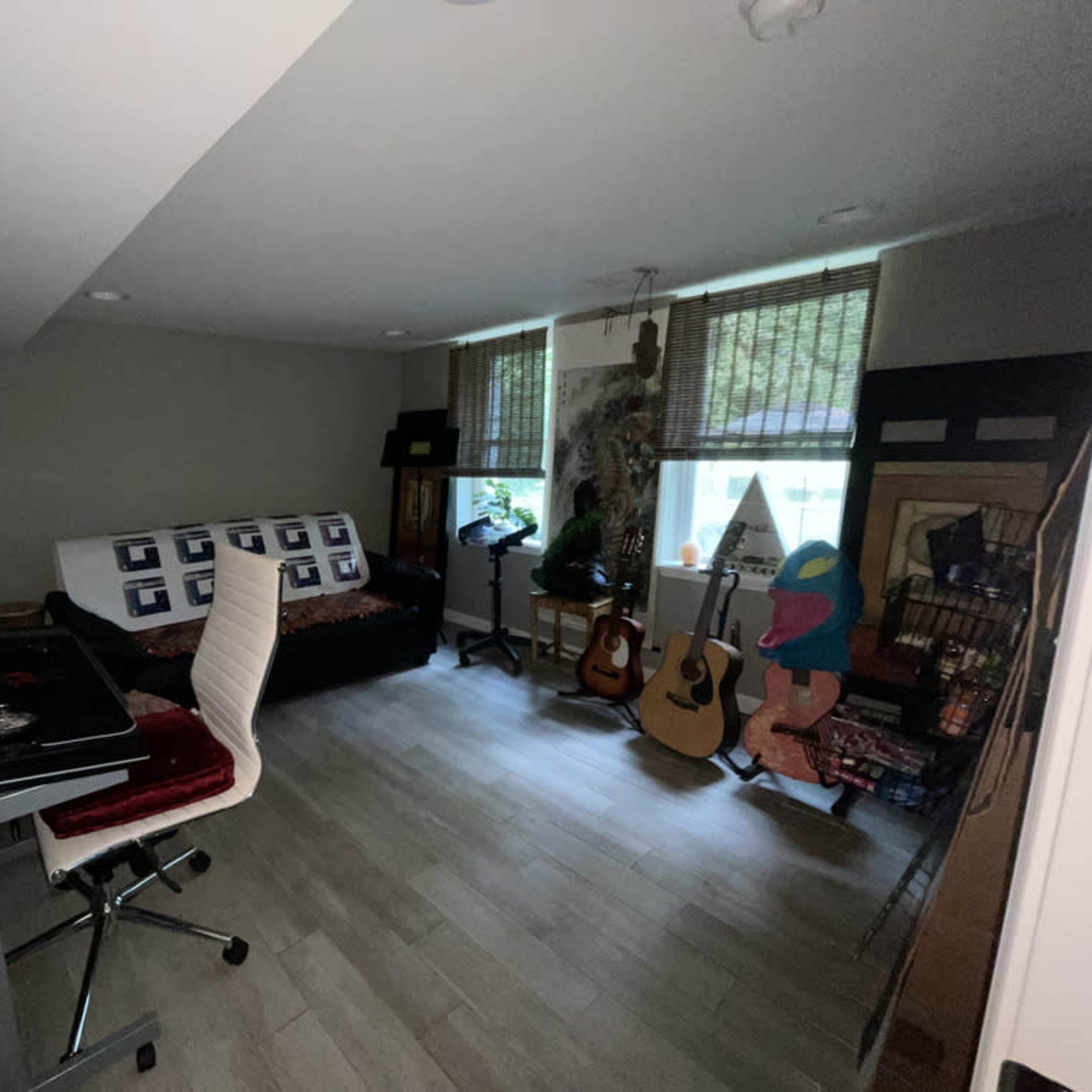 A small home office space features a desk with a chair, a couch with decorative pillows, and various musical instruments and items arranged around the room.
