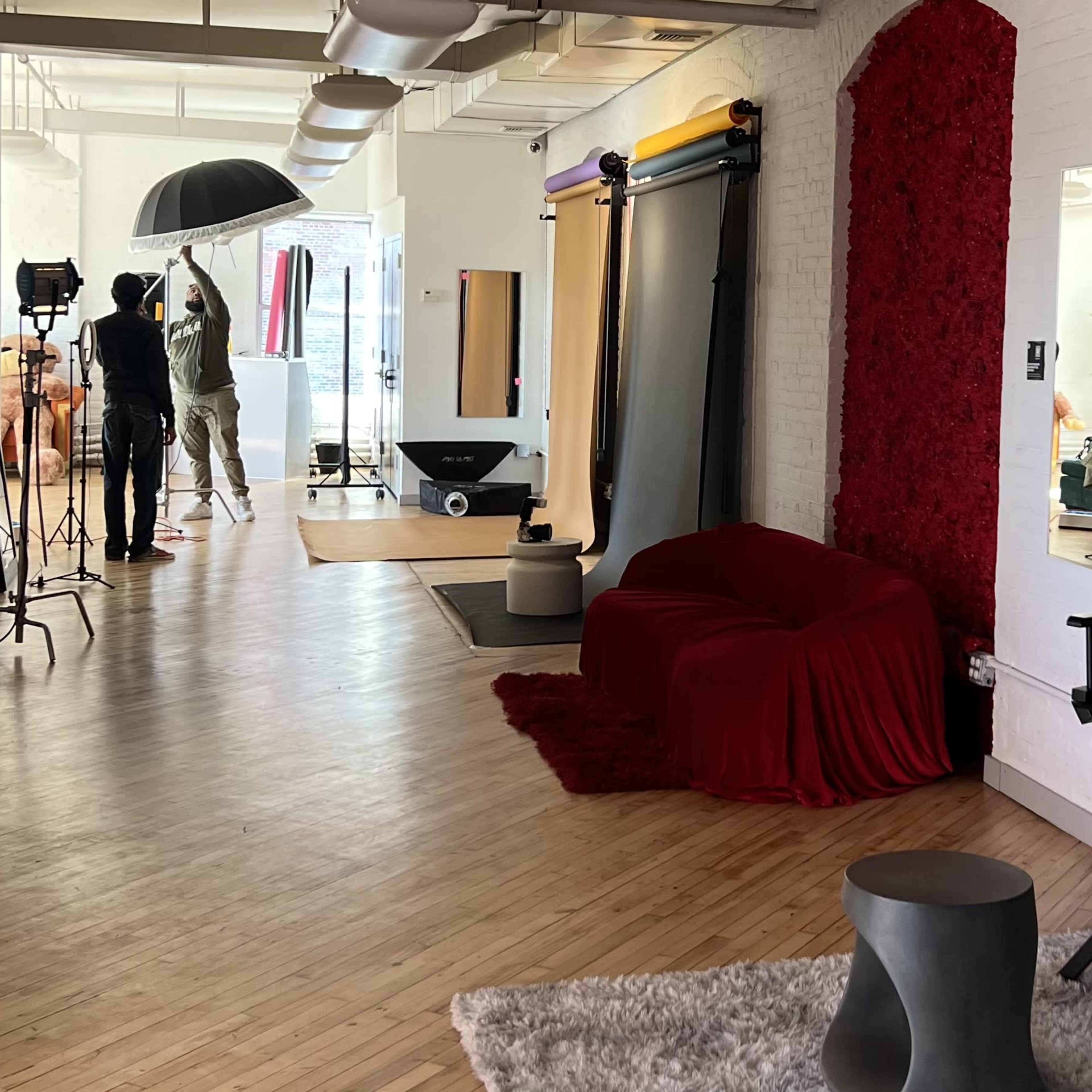 A photography studio features a red draped sofa, a grey stool, and various lighting equipment being set up in a spacious, well-lit environment.