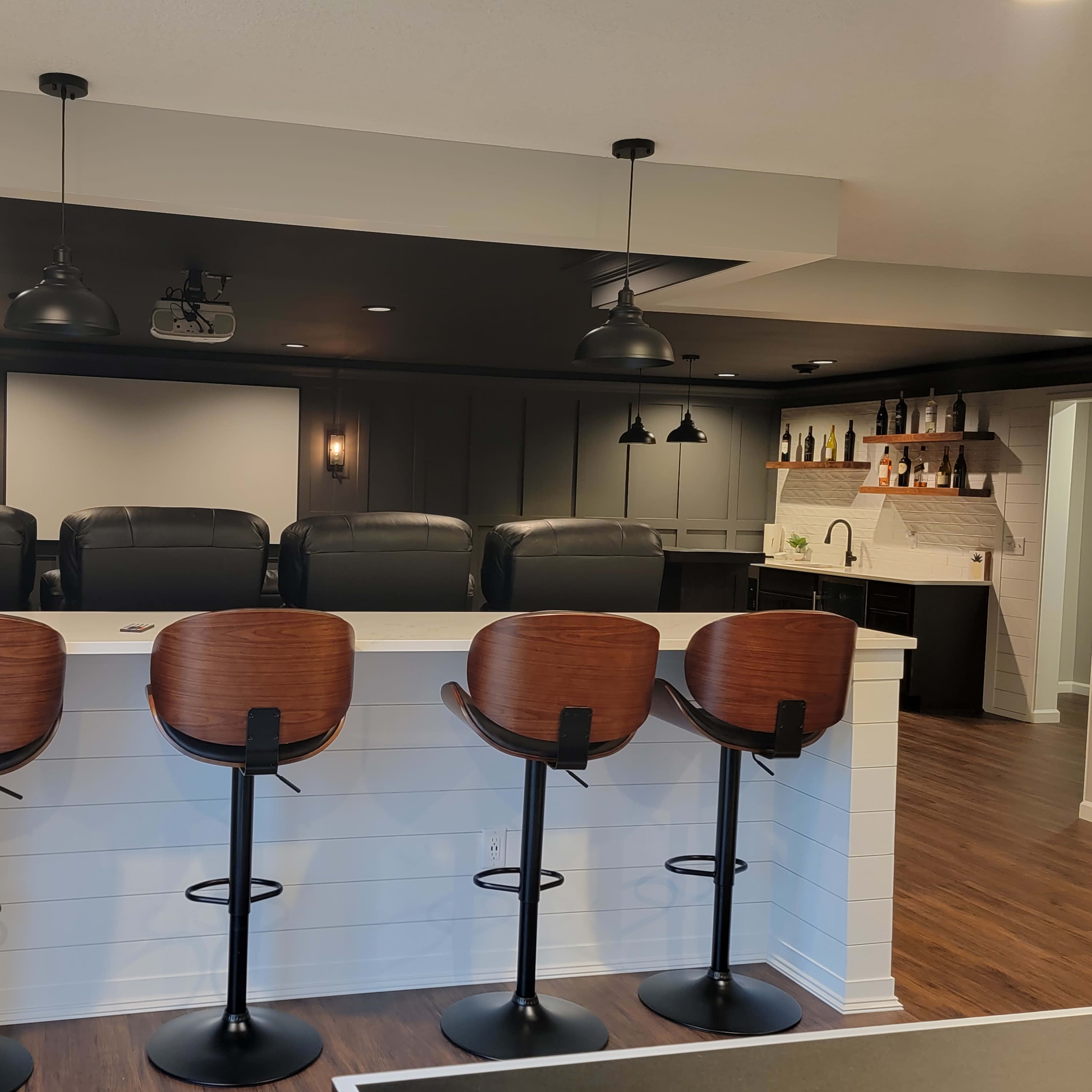 A modern home bar area with four high-backed stools lined up against a white counter, overlooking a dark-paneled entertainment space equipped with a projector screen.