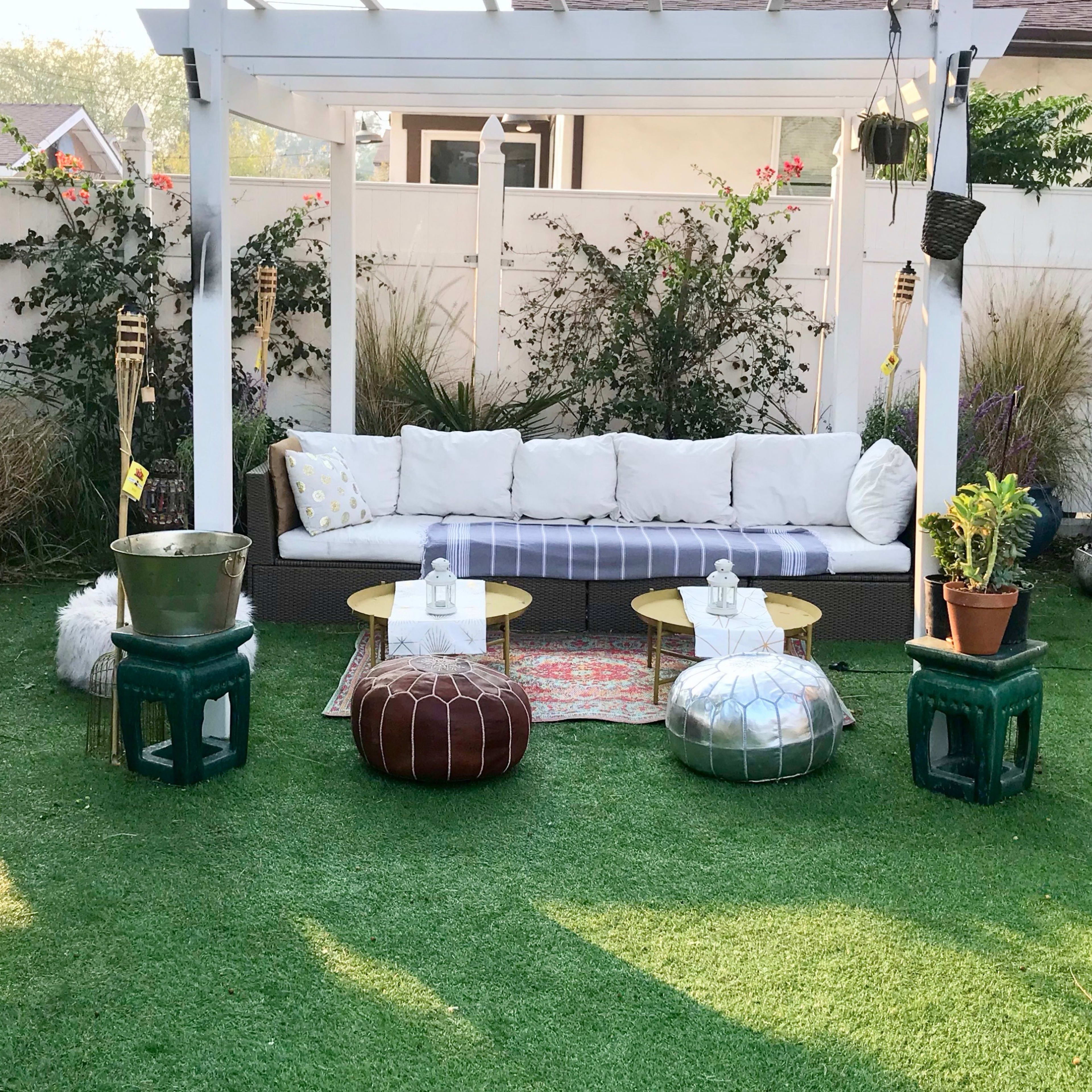 A backyard seating area with a white sofa, round tables, and decorative poufs, surrounded by green grass and plants.