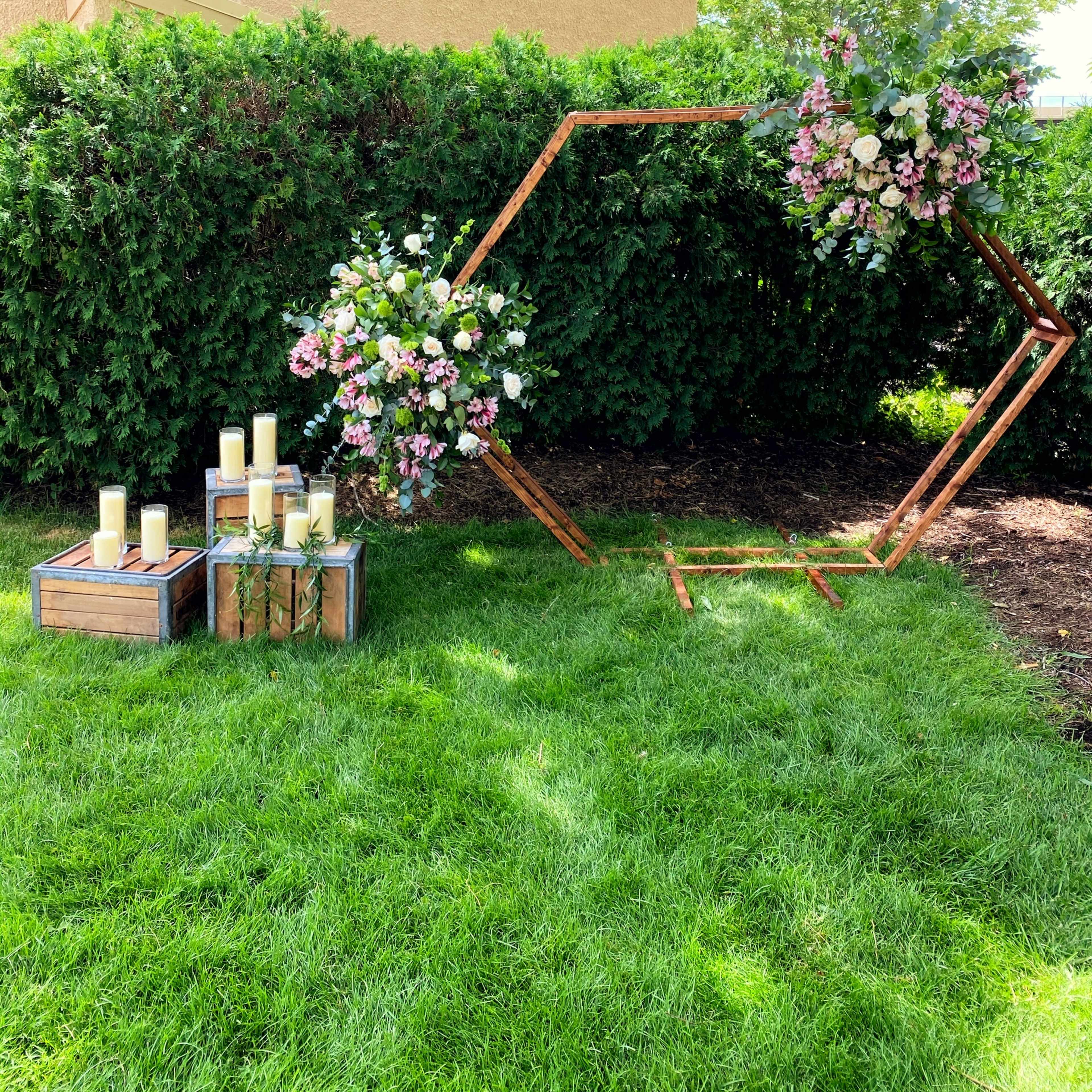 A hexagonal wooden frame adorned with colorful flowers, positioned near a cluster of candles on wooden crates, set against a backdrop of dense greenery.