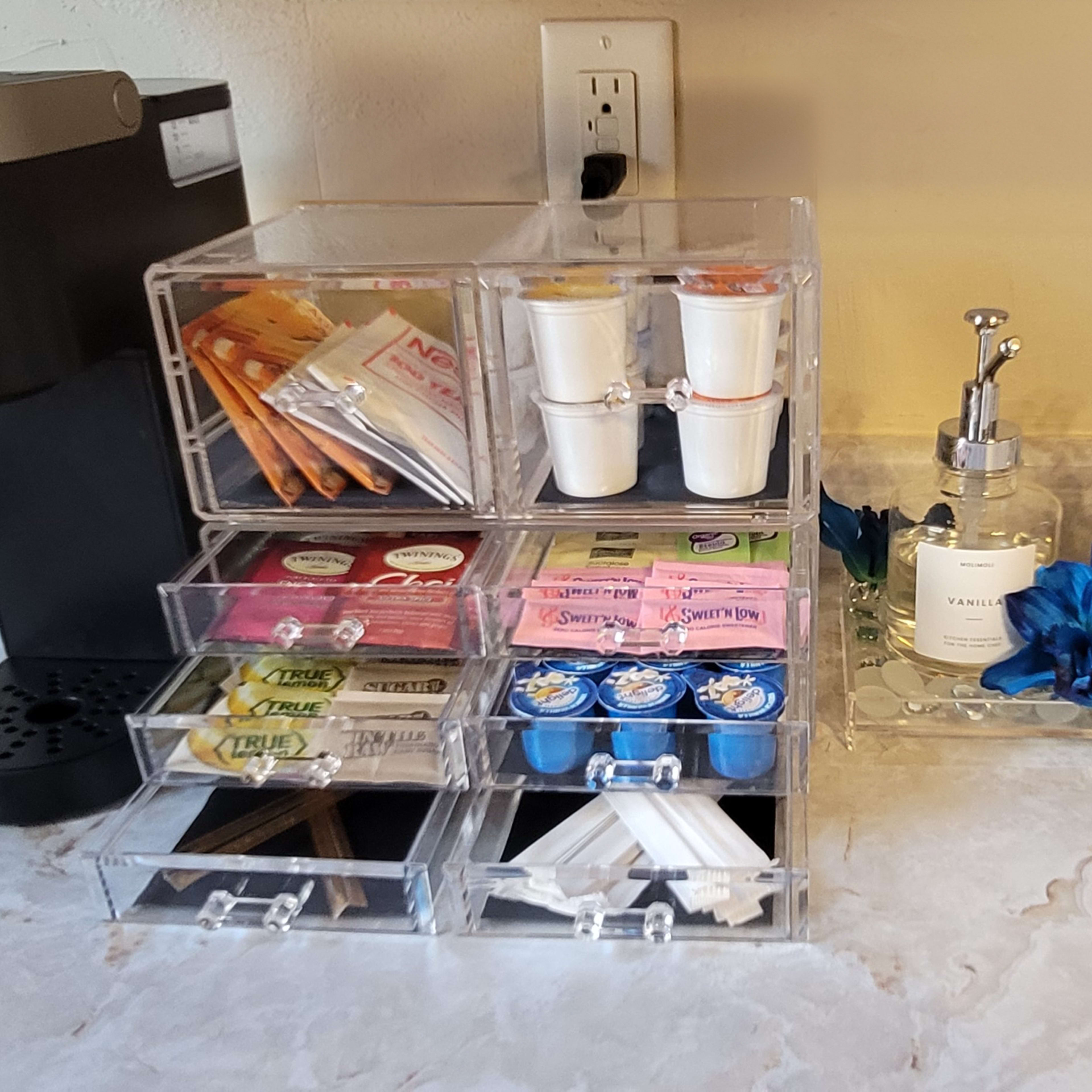 A coffee station with a Keurig machine on the left and a clear acrylic organizer containing coffee supplies and sugar packets on the right, accompanied by two glass soap dispensers.