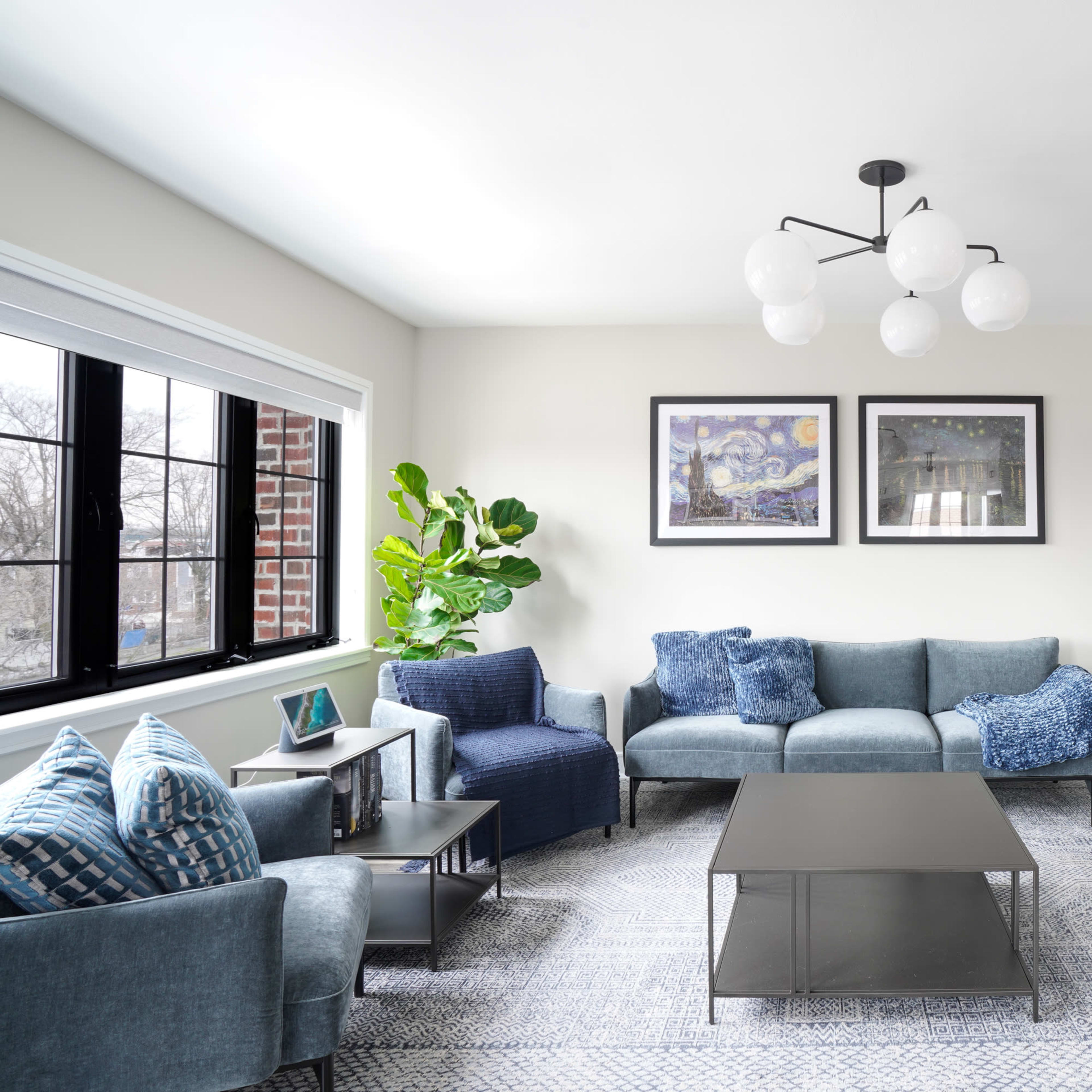 The living room features a sectional sofa, a coffee table, a large television on a stand, and two framed artworks on the wall, with a large window allowing natural light and a potted plant in the corner.