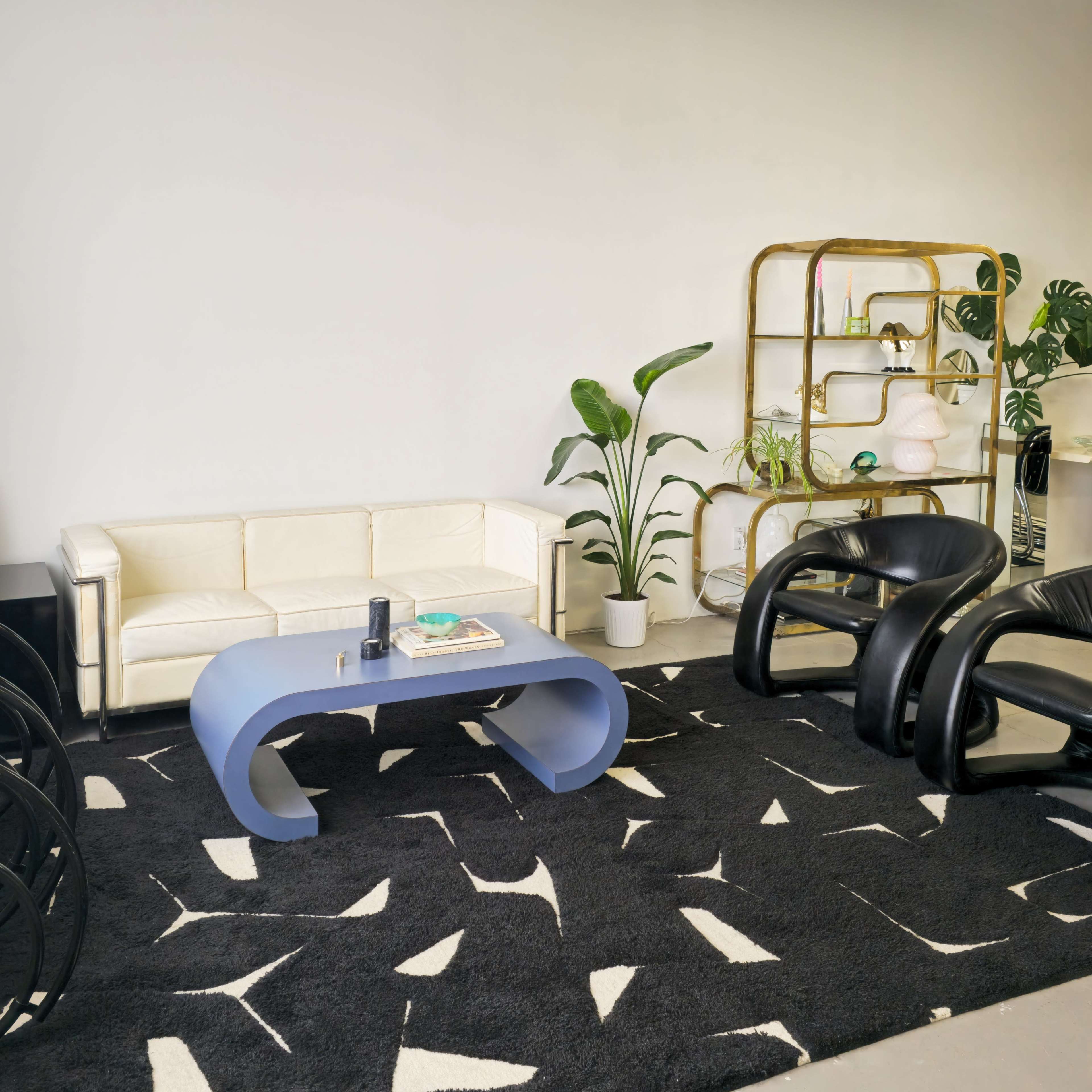 A modern living room with a white sofa, a blue coffee table, a black abstract-patterned rug, and a gold shelving unit with plants and decorative objects.
