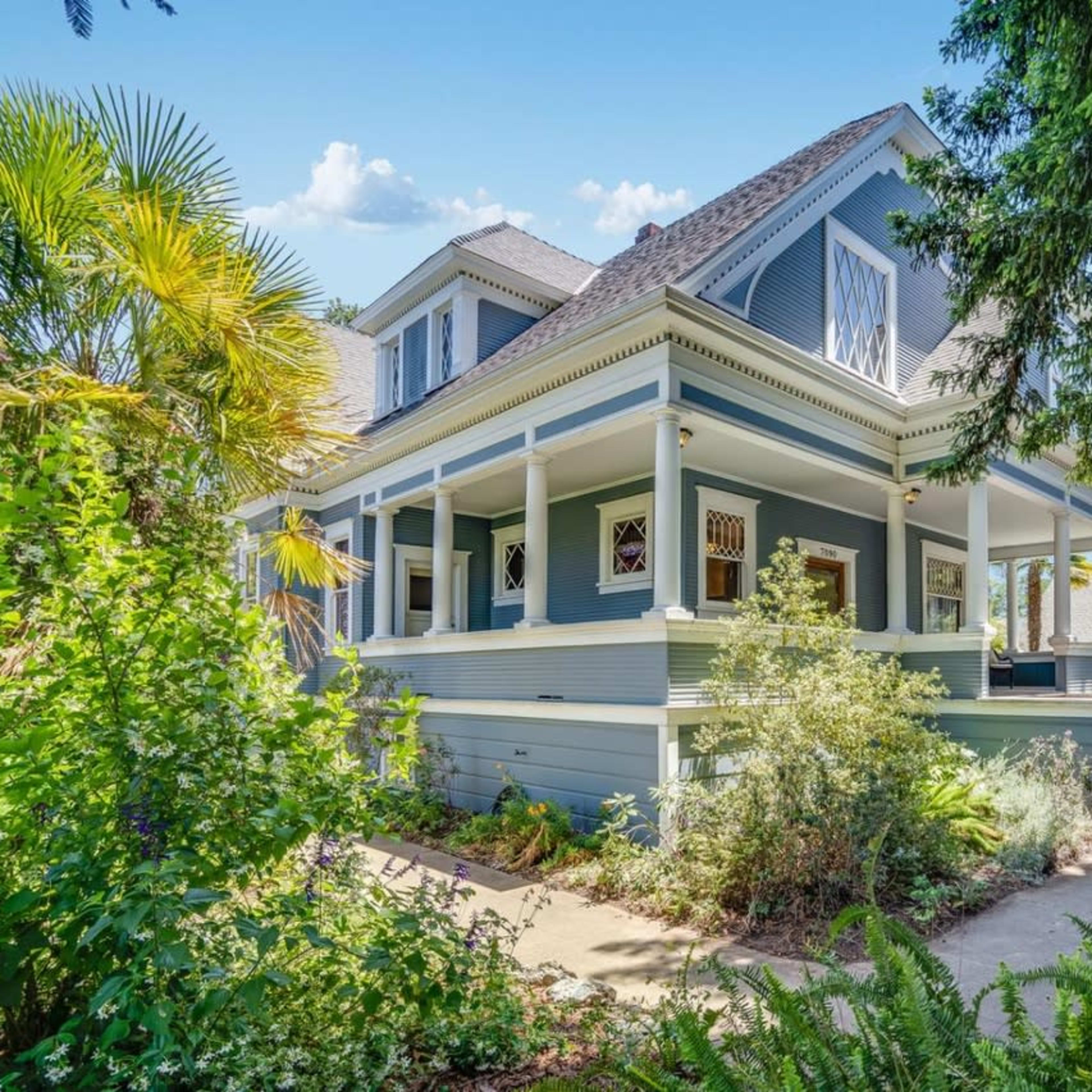 A large, two-story house with a blue exterior, surrounded by lush greenery and a well-maintained garden.