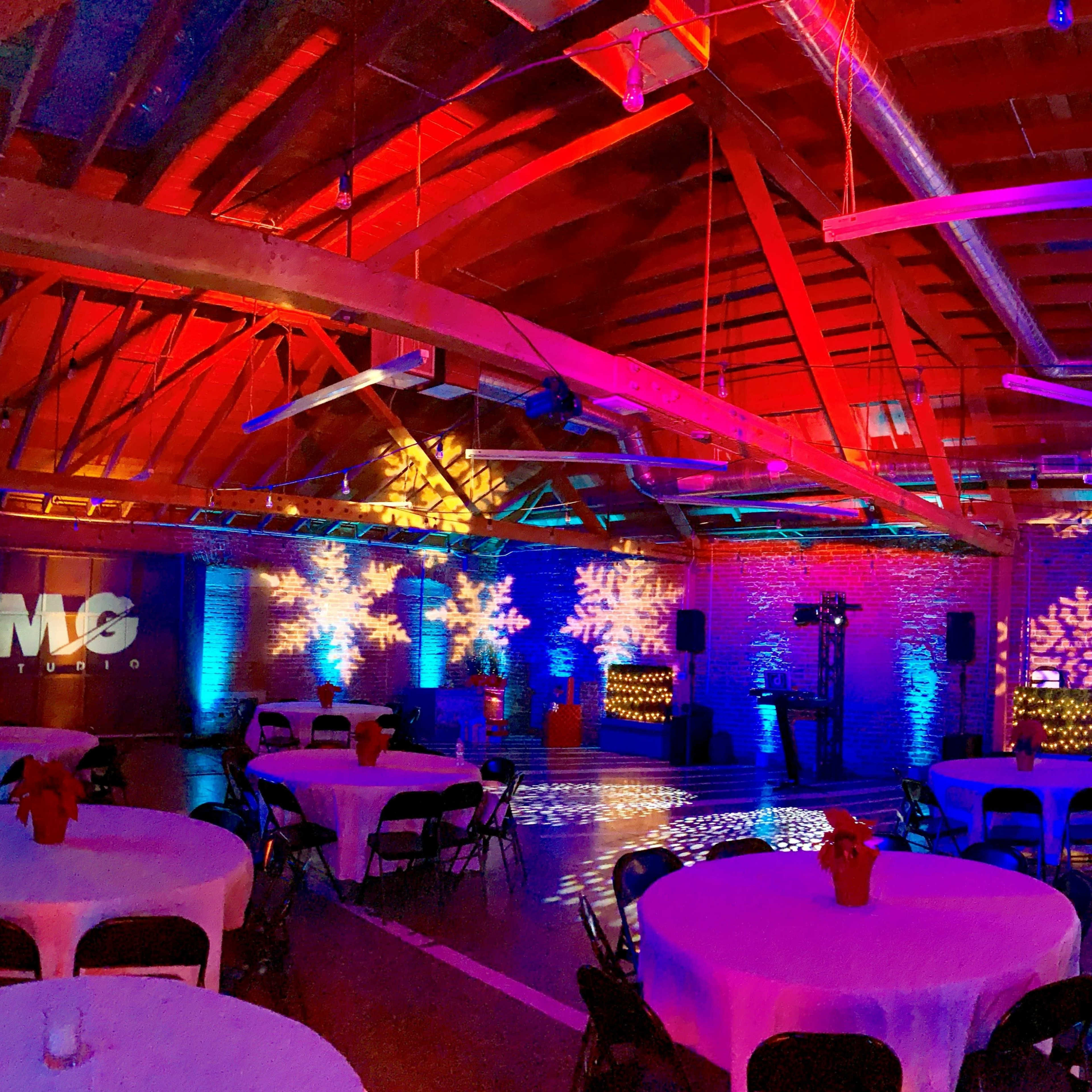 A decorated event space with tables set up under colorful lighting and a large screen displaying the letters "MG."