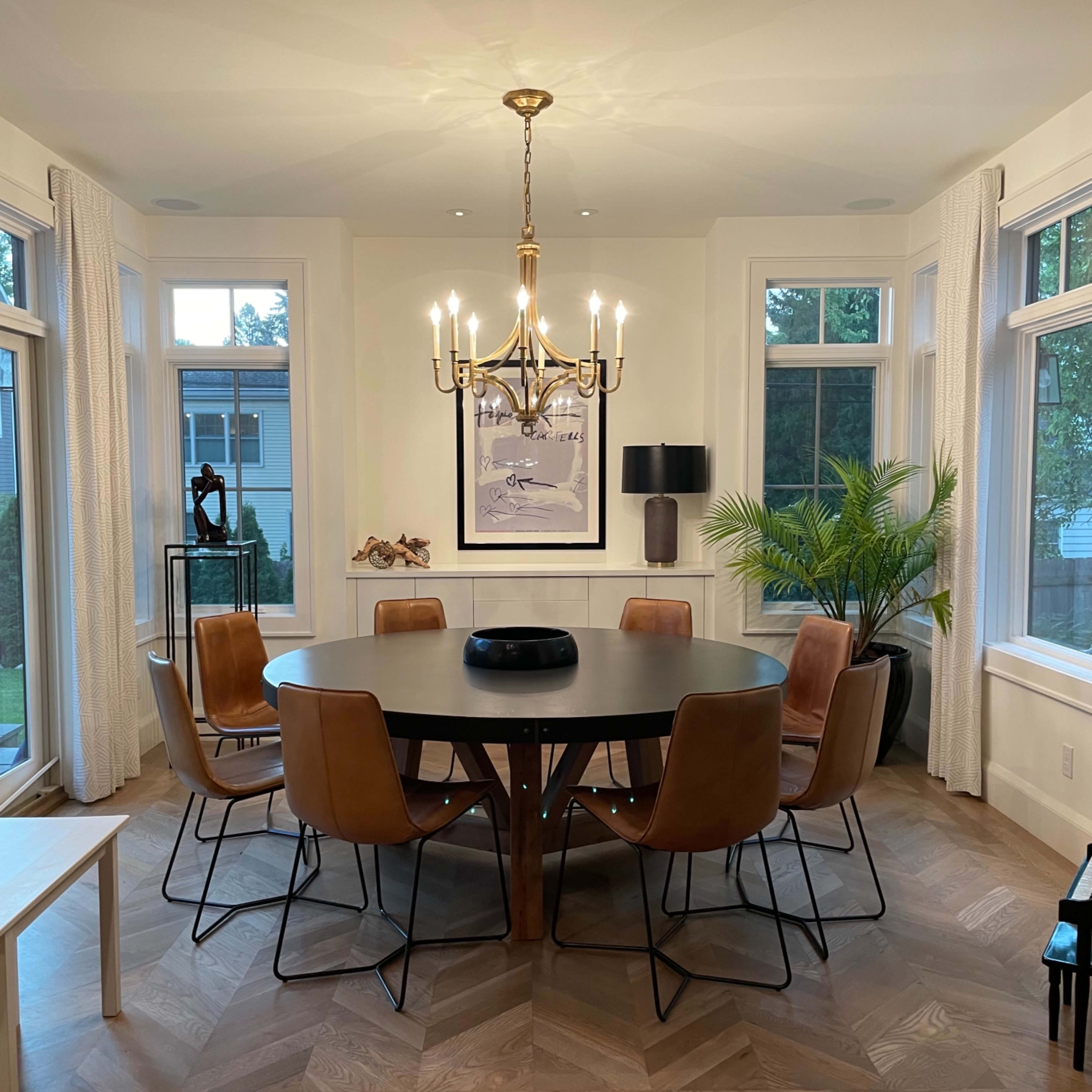 A circular dining table surrounded by leather chairs is illuminated by a chandelier in a well-lit dining room.