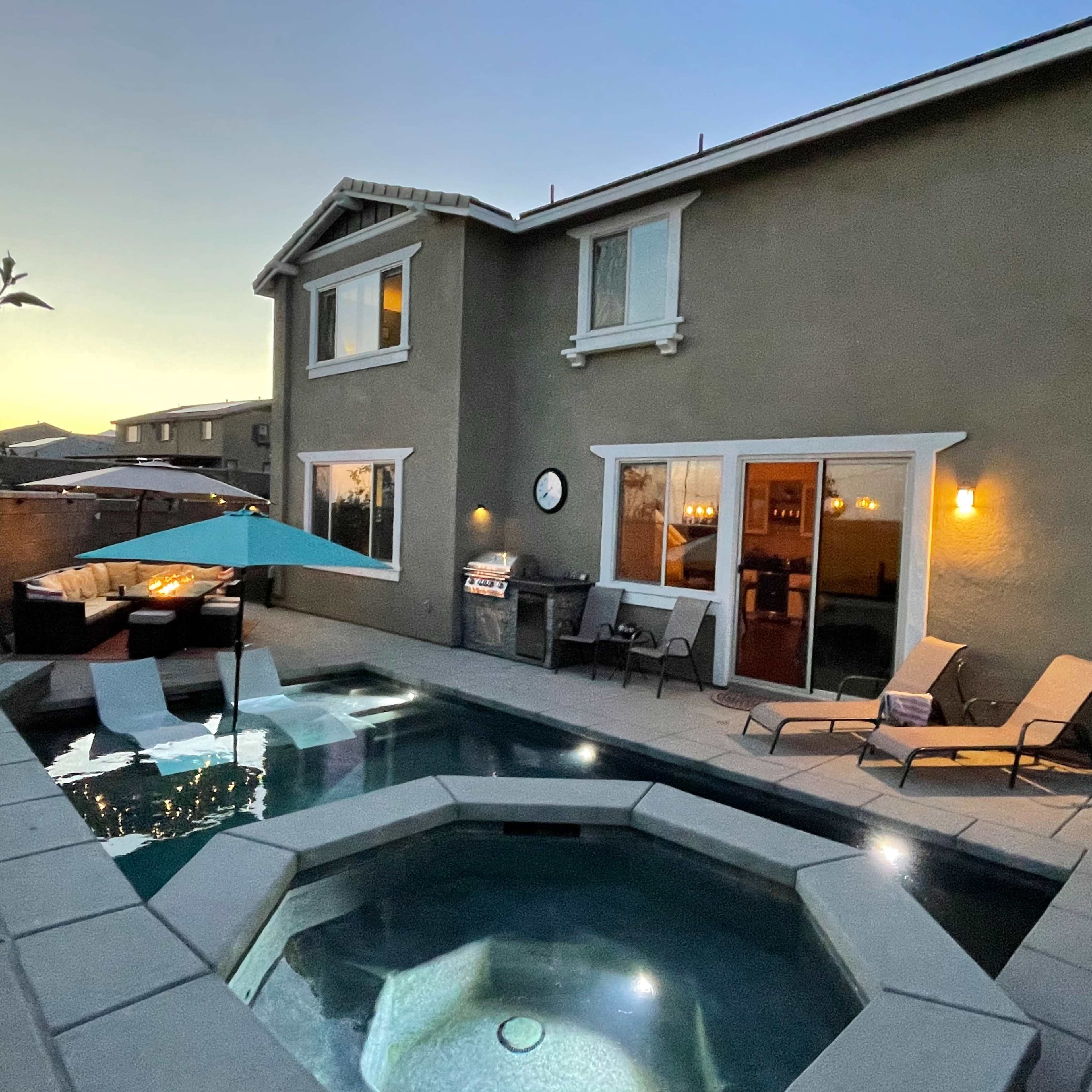 A modern home overlooking a pool area with lounge chairs, an umbrella, and a barbecue at dusk.