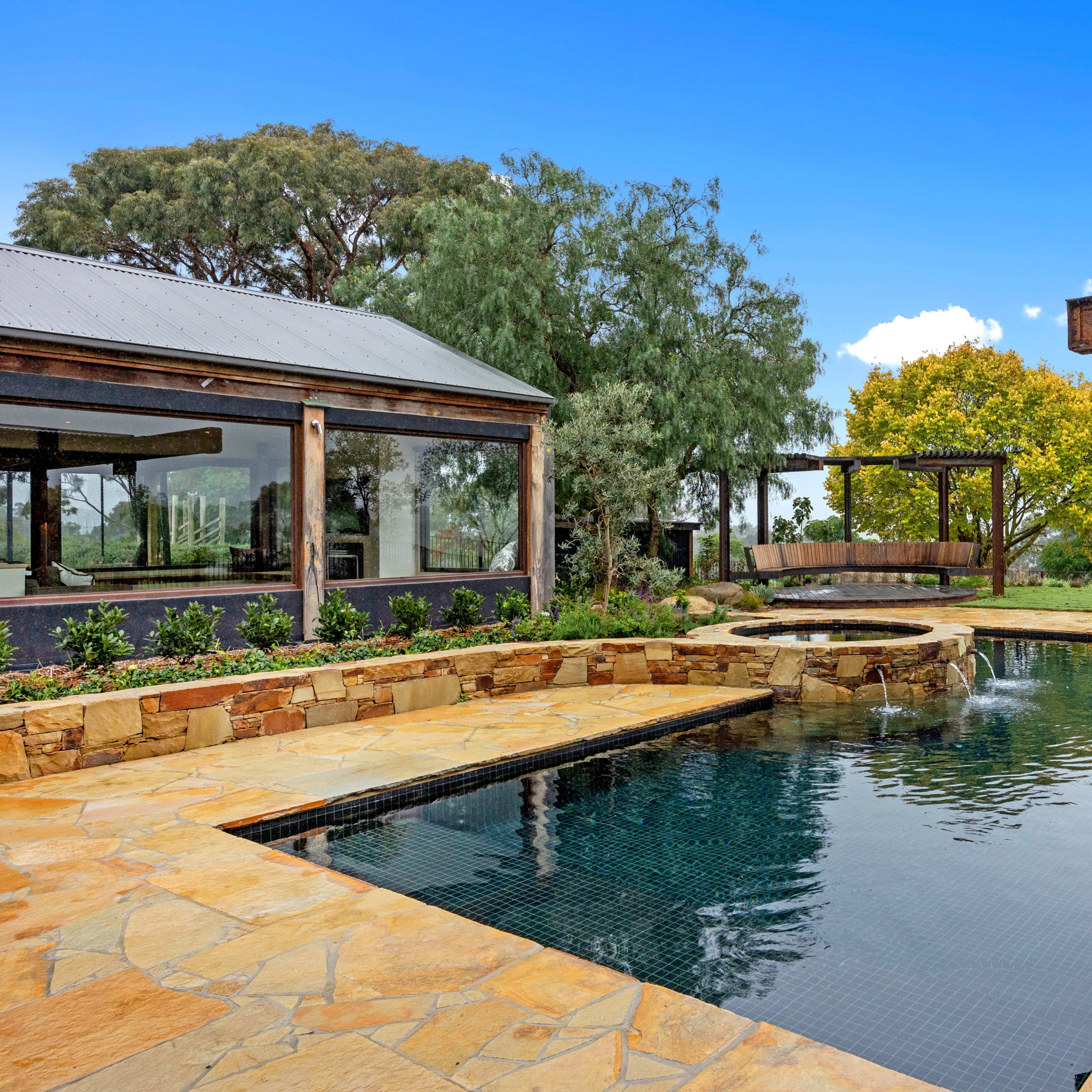 A stone patio with a swimming pool and a swing set surrounded by trees and a clear sky.