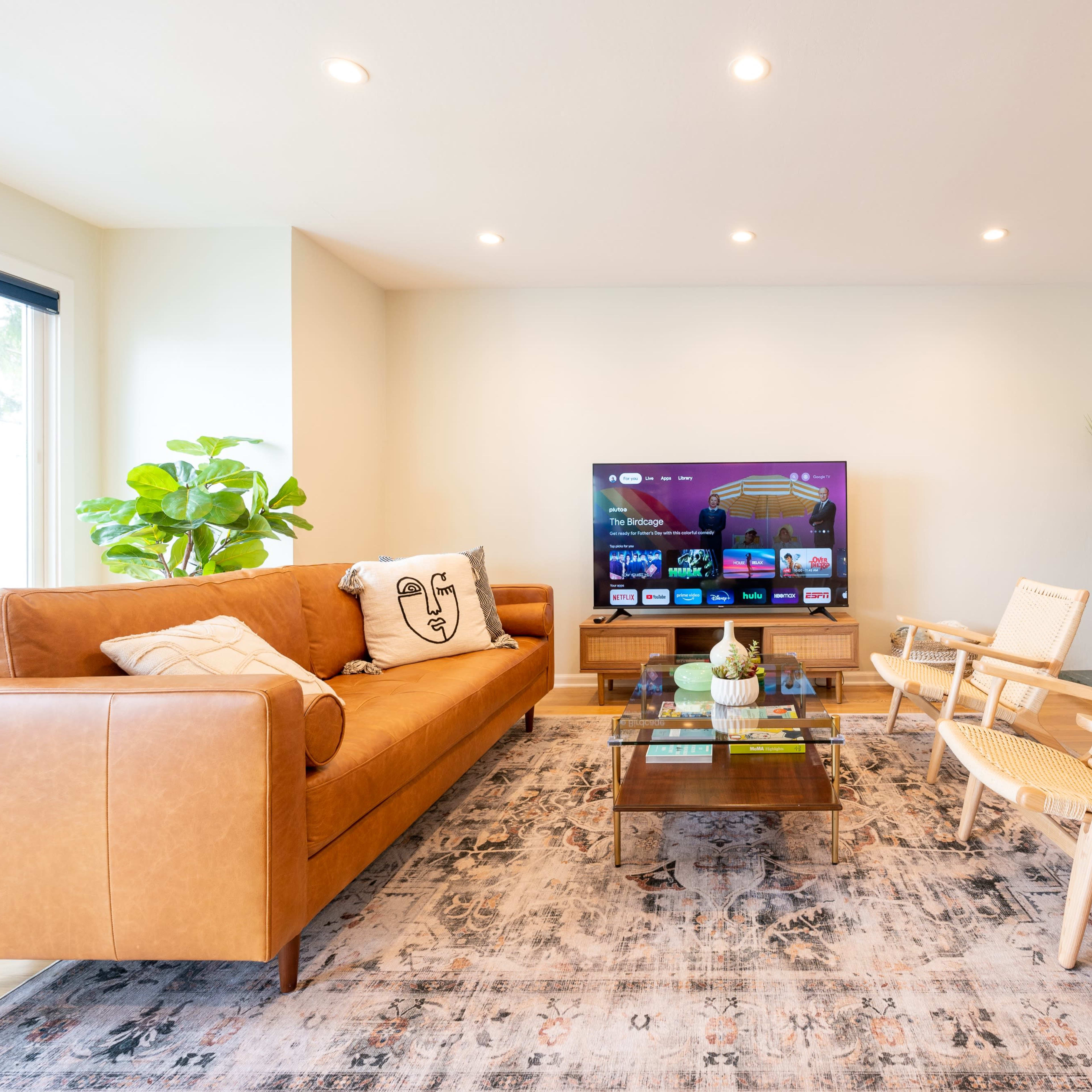 A modern living room features a leather sofa, a glass coffee table, a television on the wall, and a marble fireplace, with natural light coming from a window and a green plant in the corner.