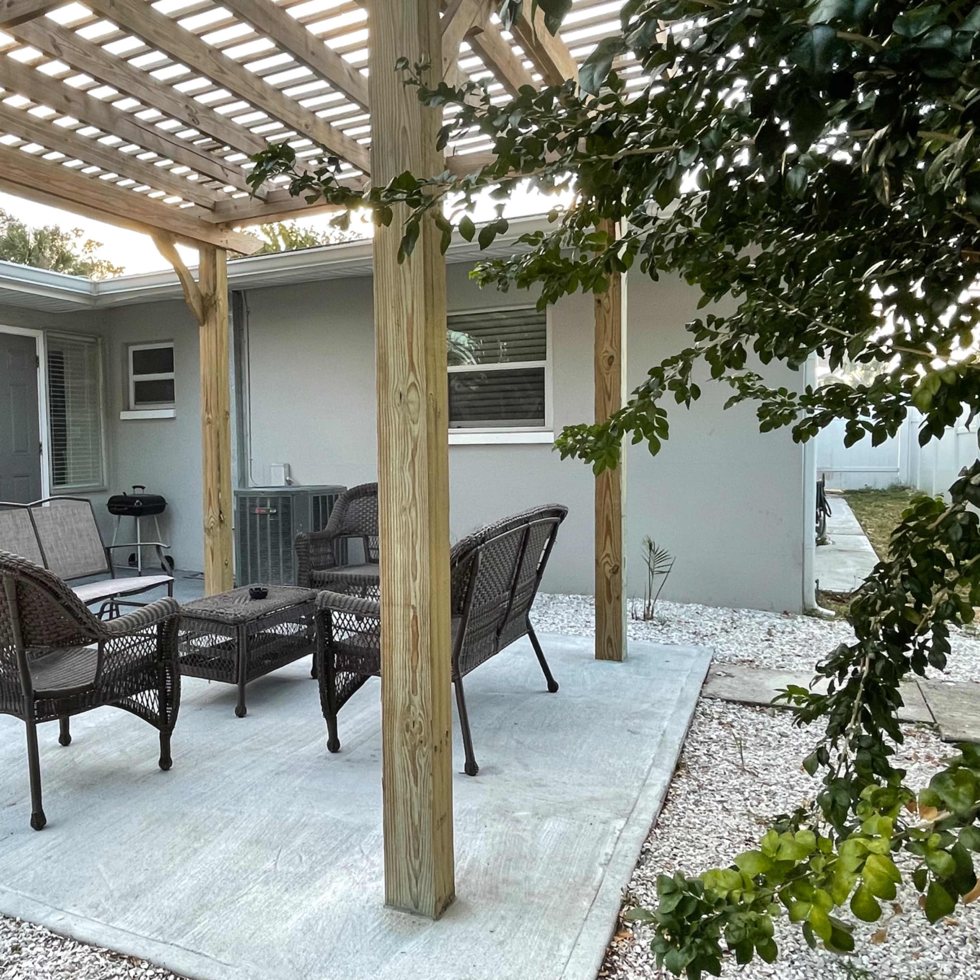 A patio area with a wooden pergola, featuring a set of four wicker chairs surrounding a rectangular table on a concrete surface.